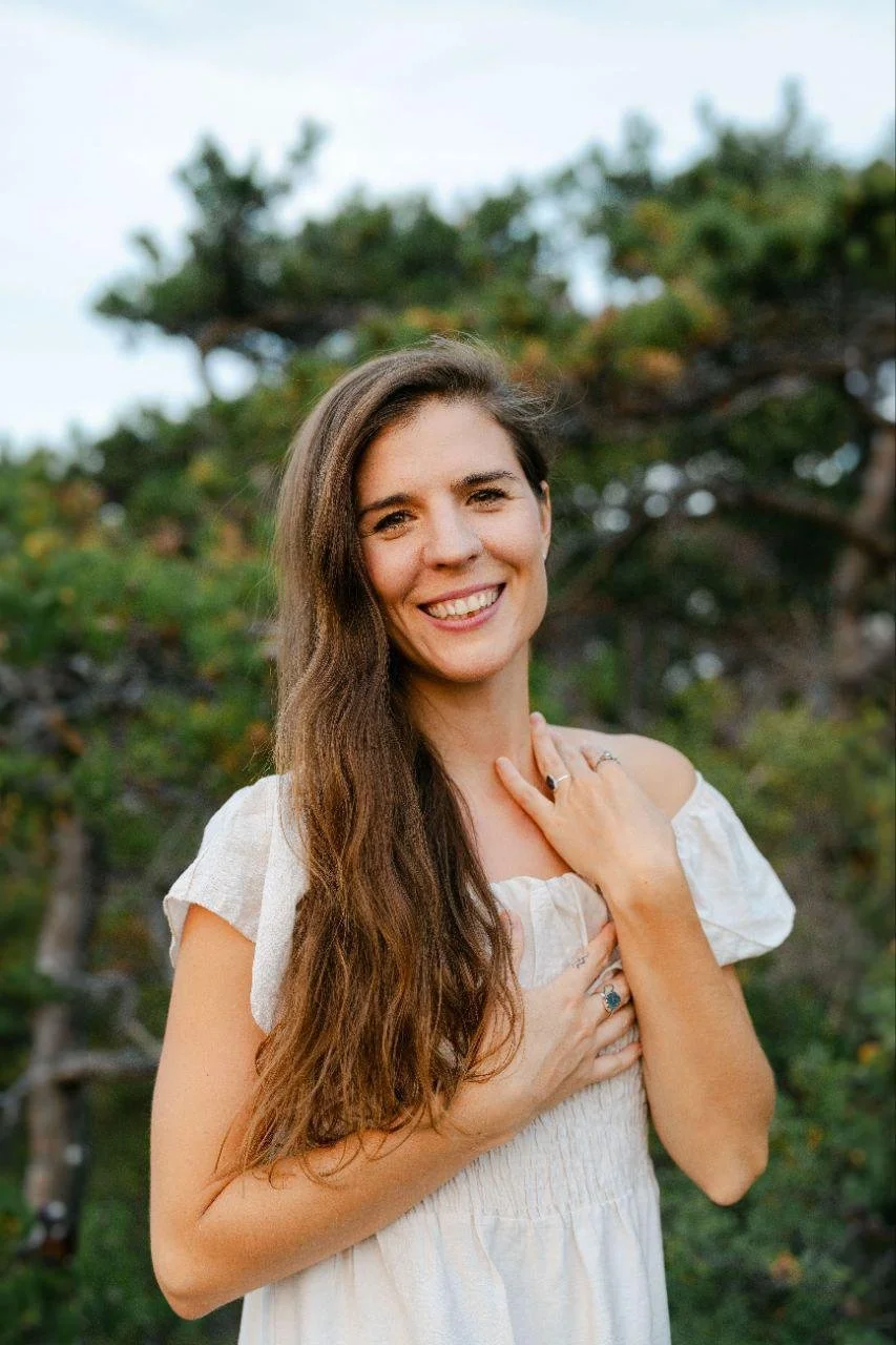 Eine lächelnde Frau mit langen braunen Haaren trägt ein weißes Kleid und steht vor einem grünen Baum, umgeben von Natur.