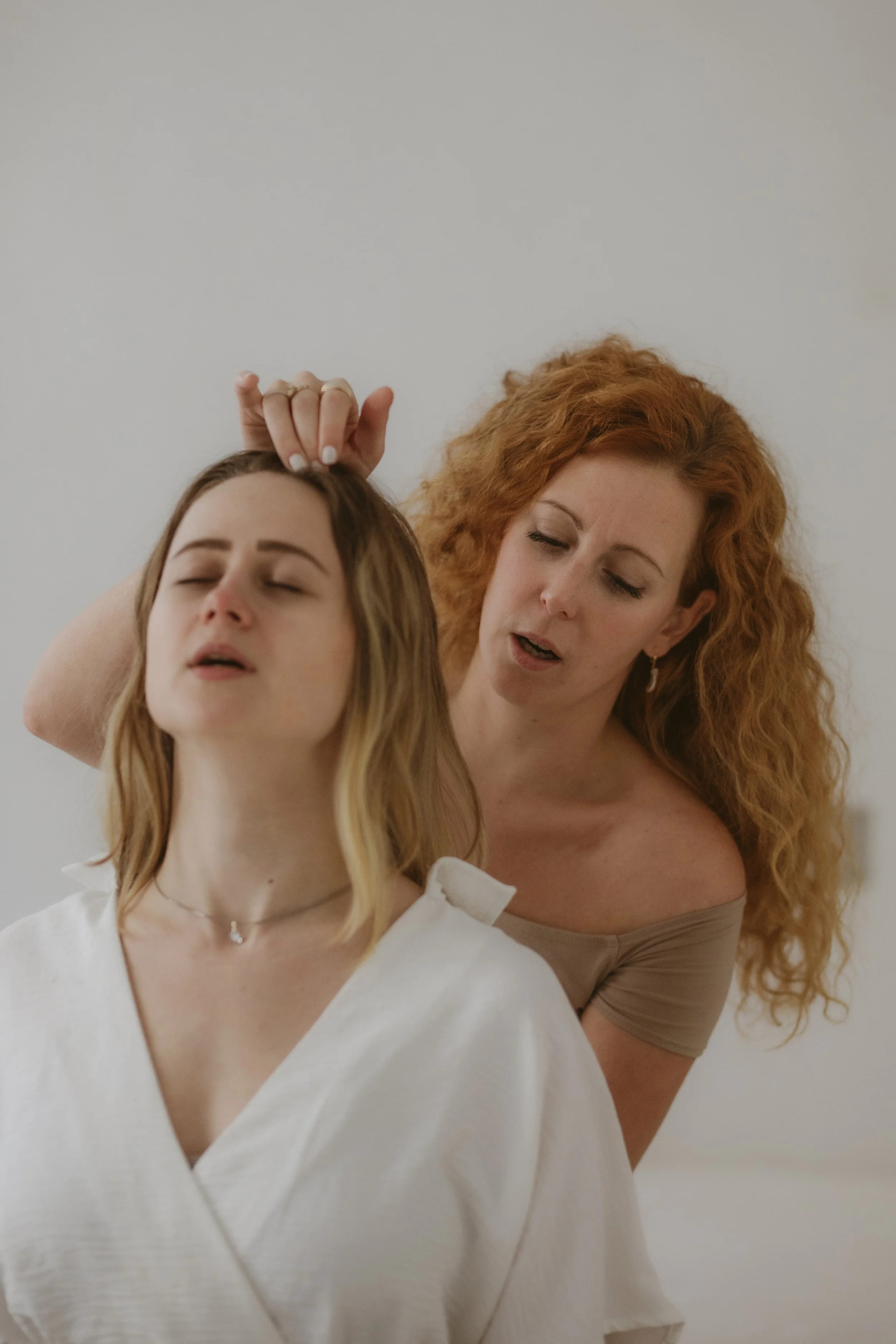 Zwei Frauen, eine mit roten, lockigen Haaren und die andere mit mittellangen, blonden Haaren, befinden sich in einer Szene, in der die ältere Frau die jüngere anscheinend streichelt oder berührt, während sie die Augen geschlossen hat.