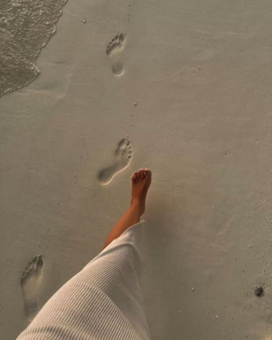 Persönlicher Aufnahmen von einem Strand mit Fußspuren im Sand, eine Person trägt weißen Stoff. Das Bild wird aus der Perspektive des Fotografen aufgenommen, der beim Gehen im Sand gefilmt wird.