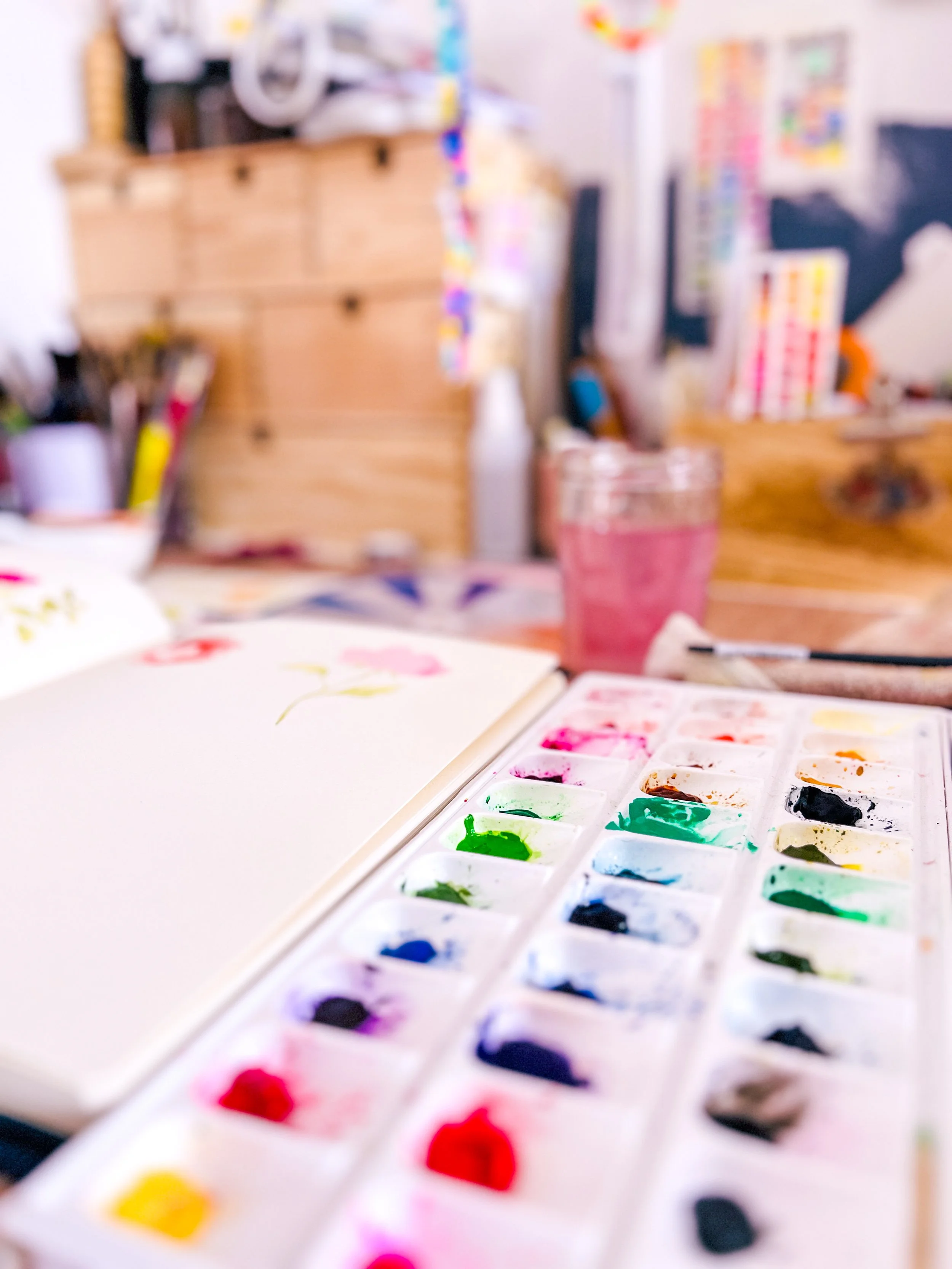 Table d'art avec palette de peinture aquarelle colorée, un carnet de croquis avec un dessin floral, et un pinceau. Arrière-plan flou d'un espace de travail artistique.
