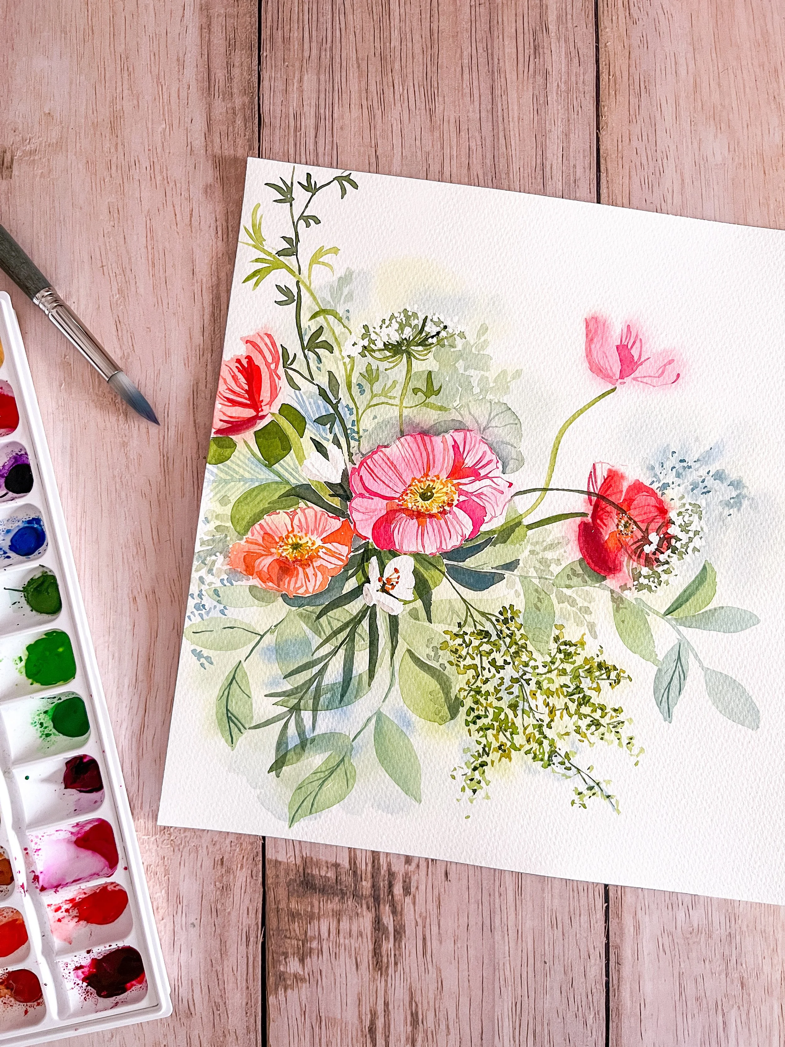 Une peinture aquarelle de fleurs colorées sur du papier blanc, avec un pinceau à côté et une palette de peinture à gauche, le tout posé sur une surface en bois clair.
