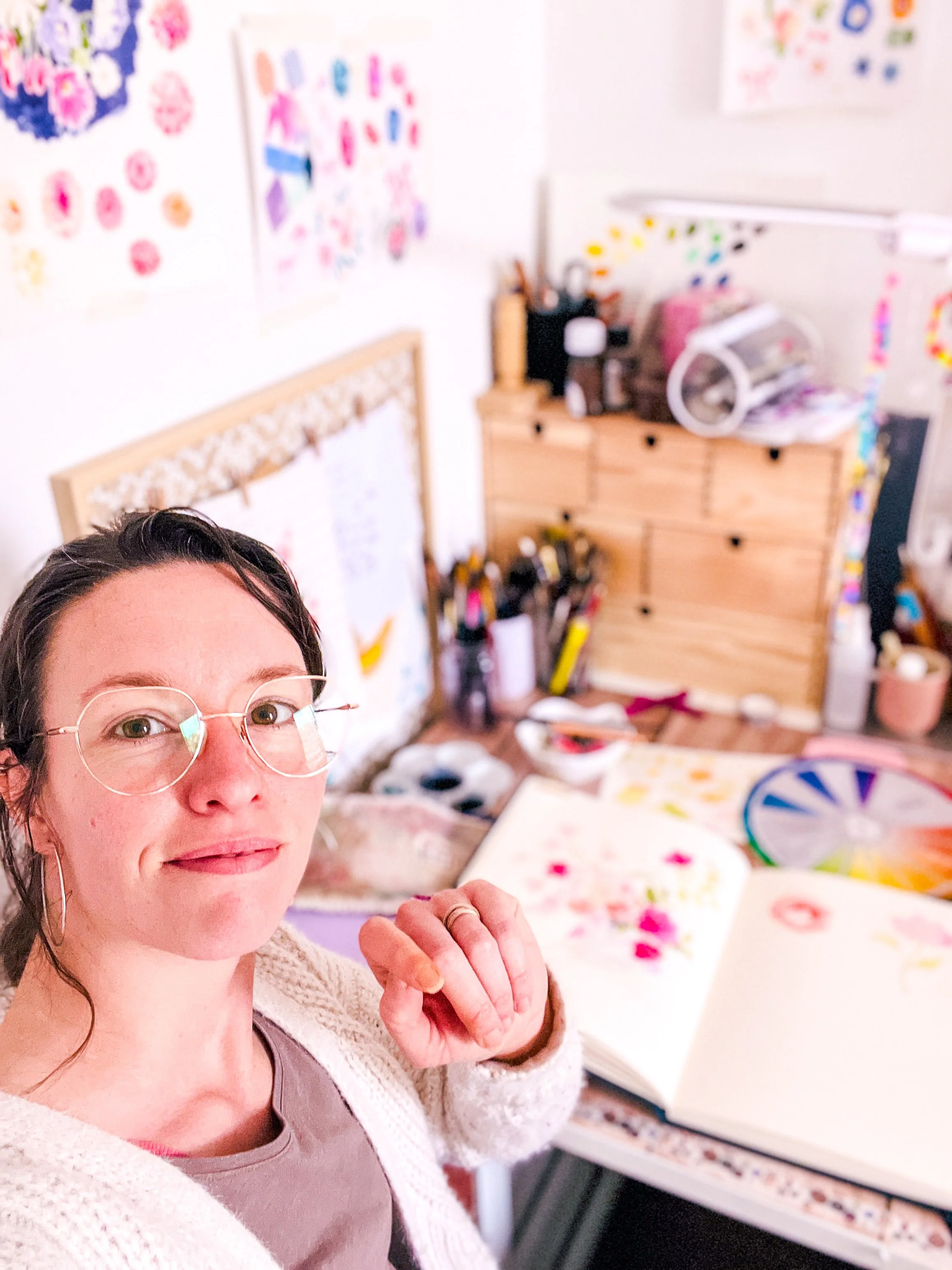 Une femme avec des lunettes prend un selfie dans un atelier d'art avec des peintures, des pinceaux, un cercle de roue de couleurs et des dessins colorés sur les murs.