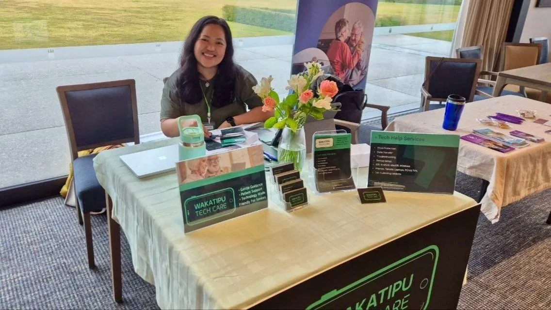 A woman sitting at a tech support booth with a laptop, flowers, and info signs, indoors with large glass windows showing a grassy area outside.