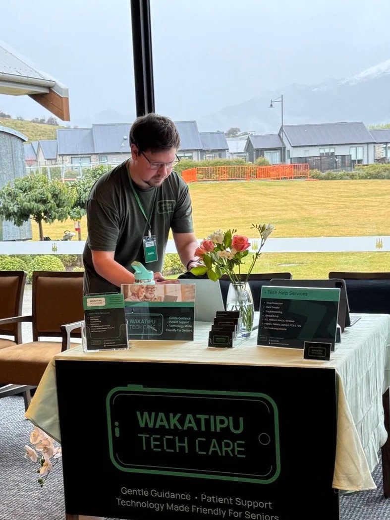 A man attends a tech care event at a table with informational signs and flowers in a glass vase, with a large window showing an outdoor landscape of houses and mountains in the background.