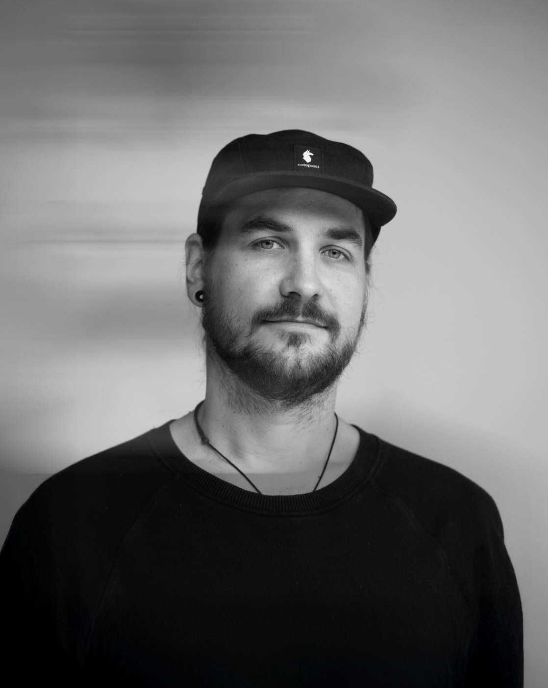 A black-and-white portrait of a man with a beard, wearing a cap and a black shirt, looking at the camera with a neutral expression.