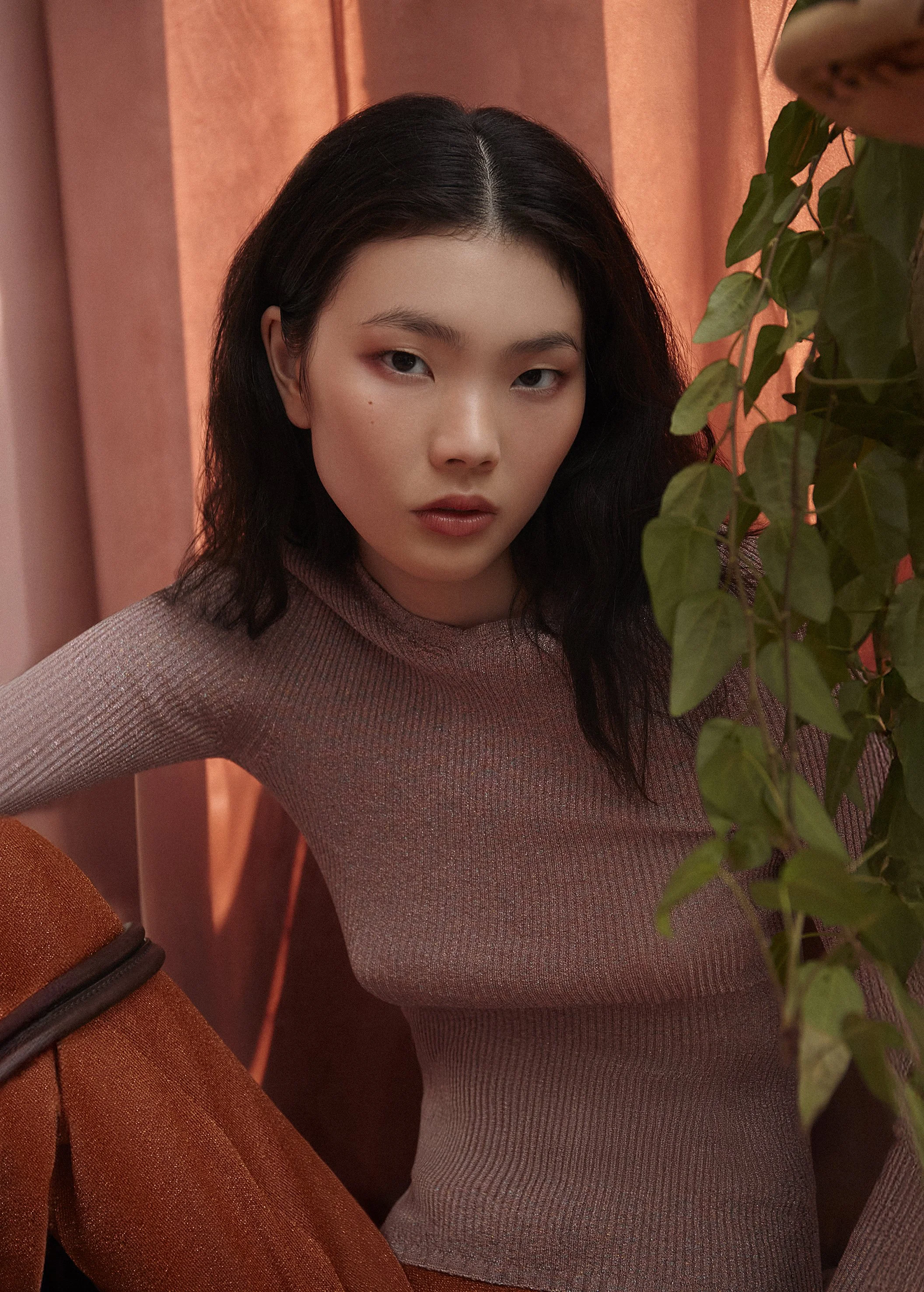 Young woman with dark wavy hair wearing a light pink ribbed turtleneck sweater, posing in front of pink curtains with a green potted plant partially covering her. The lighting is warm, creating a soft, intimate atmosphere.