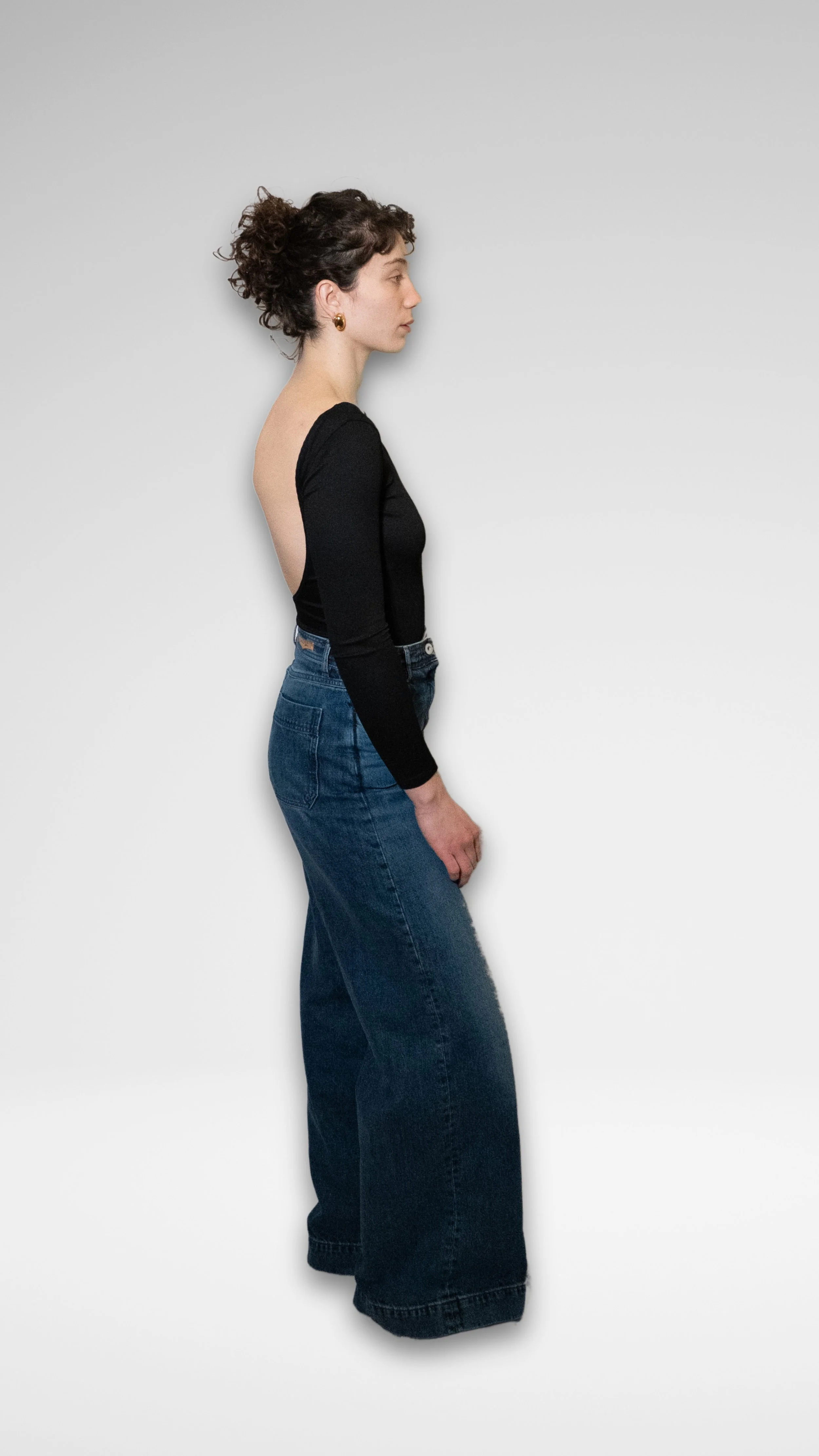 Femme en profil, portant un haut noir à dos nu et un jean taille haute amples, avec des boucles d'oreilles, devant un mur blanc.