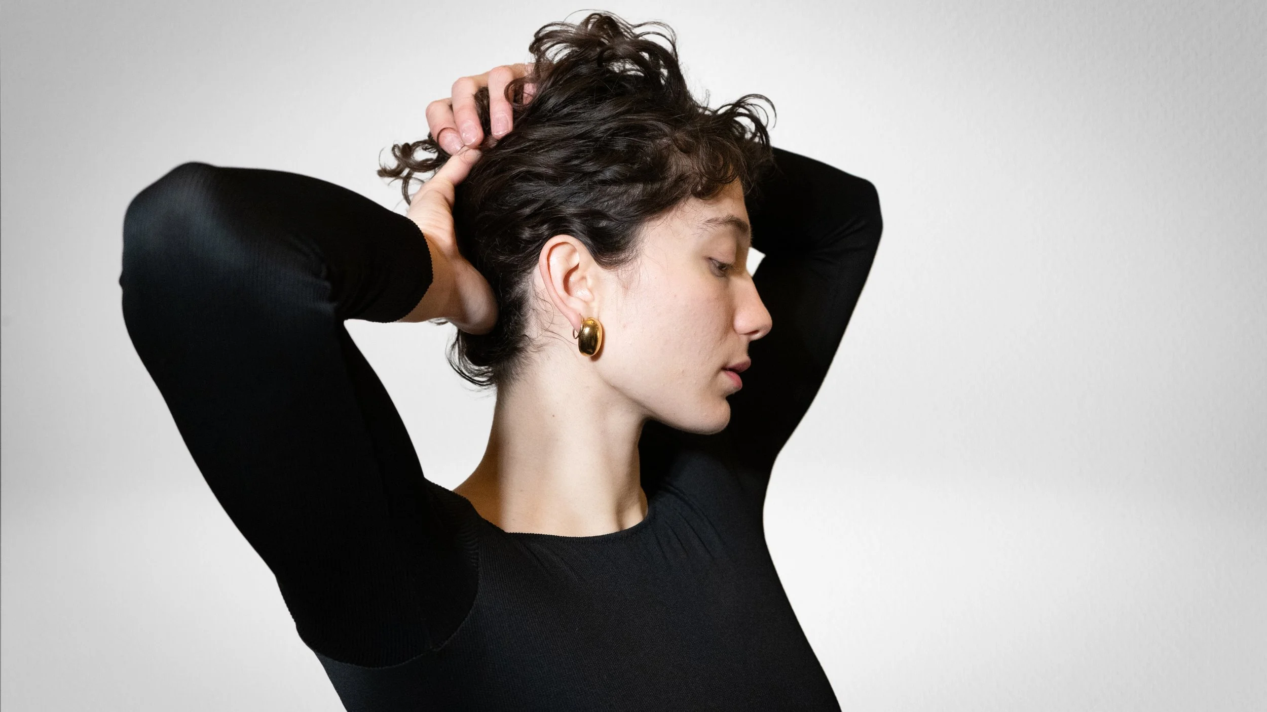 Jeune femme avec boucle d'oreille dorée, portant un haut noir, les bras levés derrière la tête, regardant vers la droite, contre un fond gris clair.