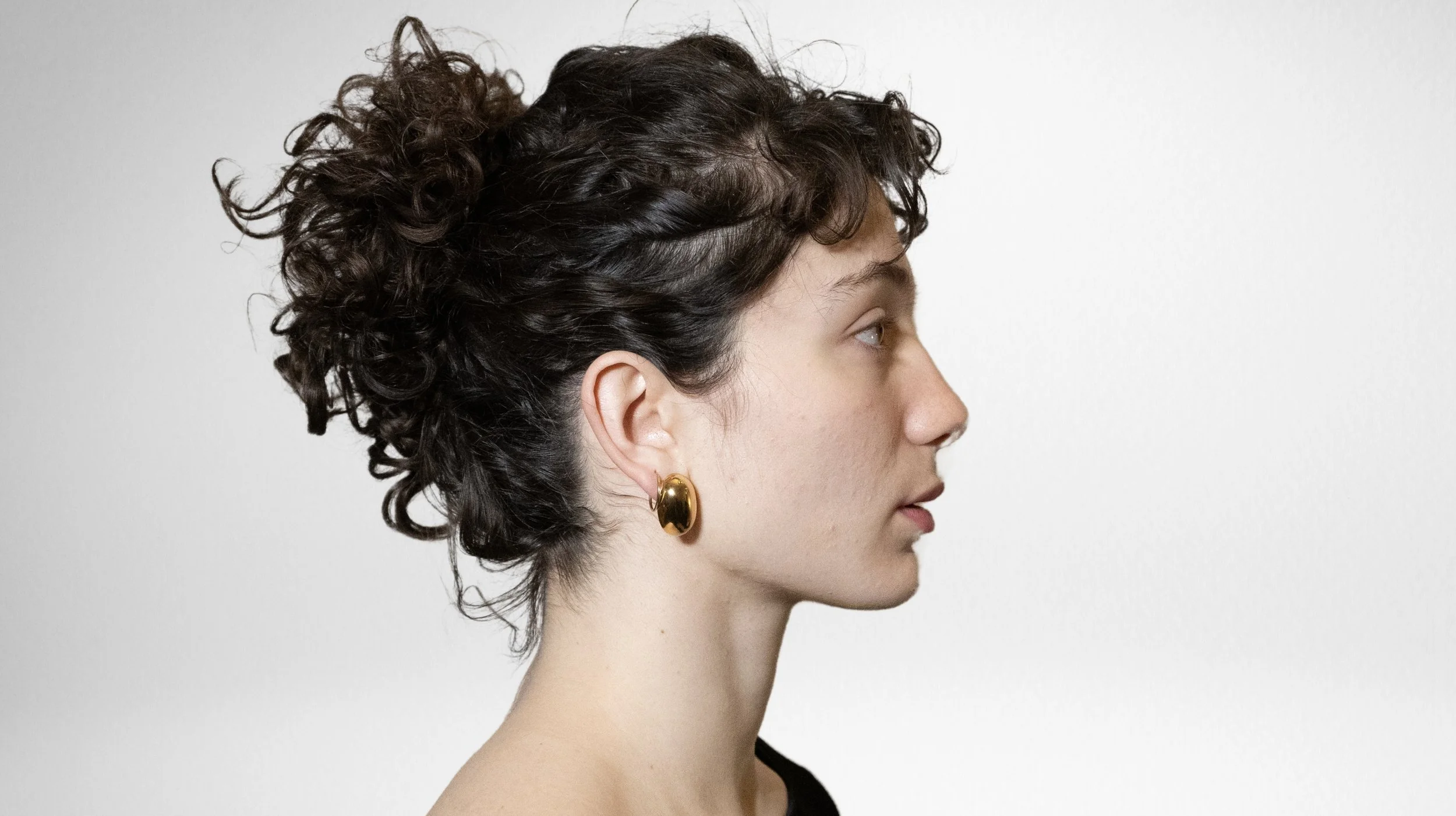 Profil d'une jeune femme avec cheveux bouclés attachés en chignon, portant une boucle d'oreille en or, sur fond blanc.