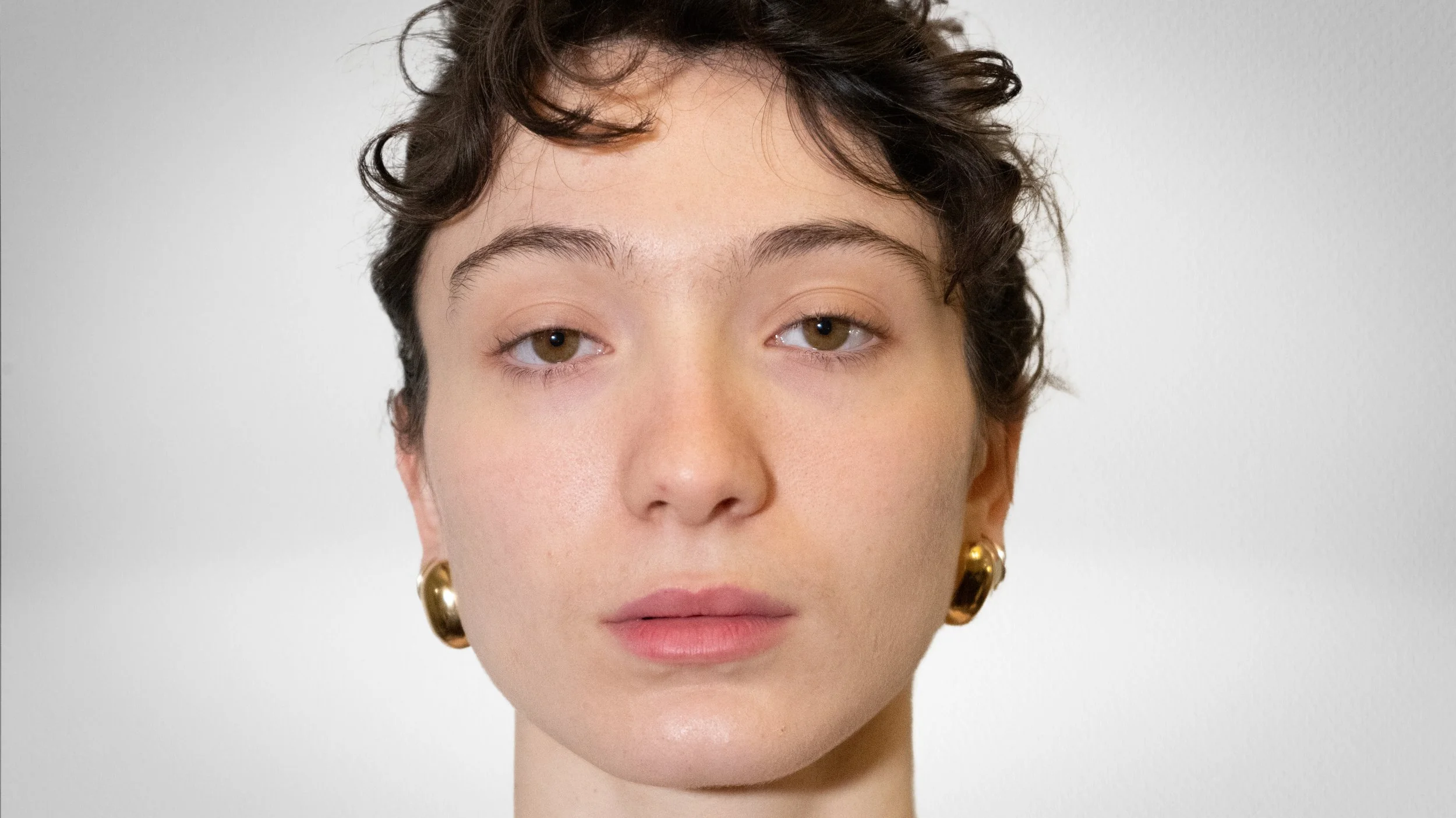 Portrait d'une jeune femme avec des cheveux courts et bouclés, portant des boucles d'oreilles dorées, avec un fond blanc