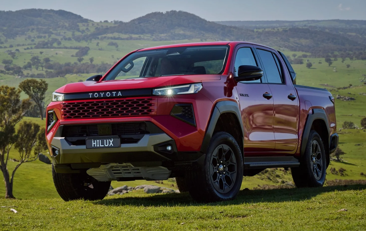 Red Toyota Hilux pickup truck parked on a grassy field with rolling hills and trees in the background.