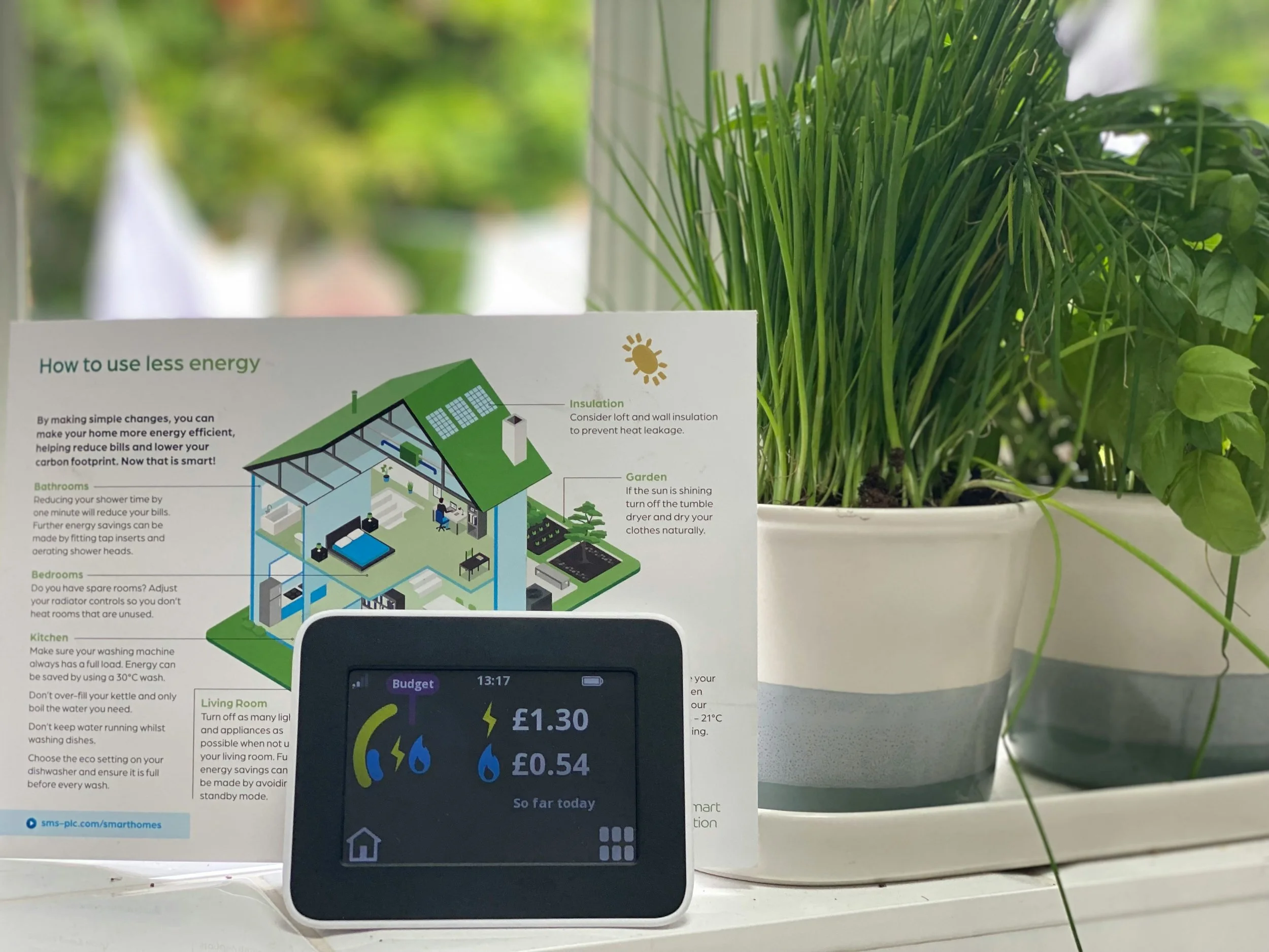 A digital energy monitor, placed in front of an illustrated home energy efficiency guide and a potted green plant.