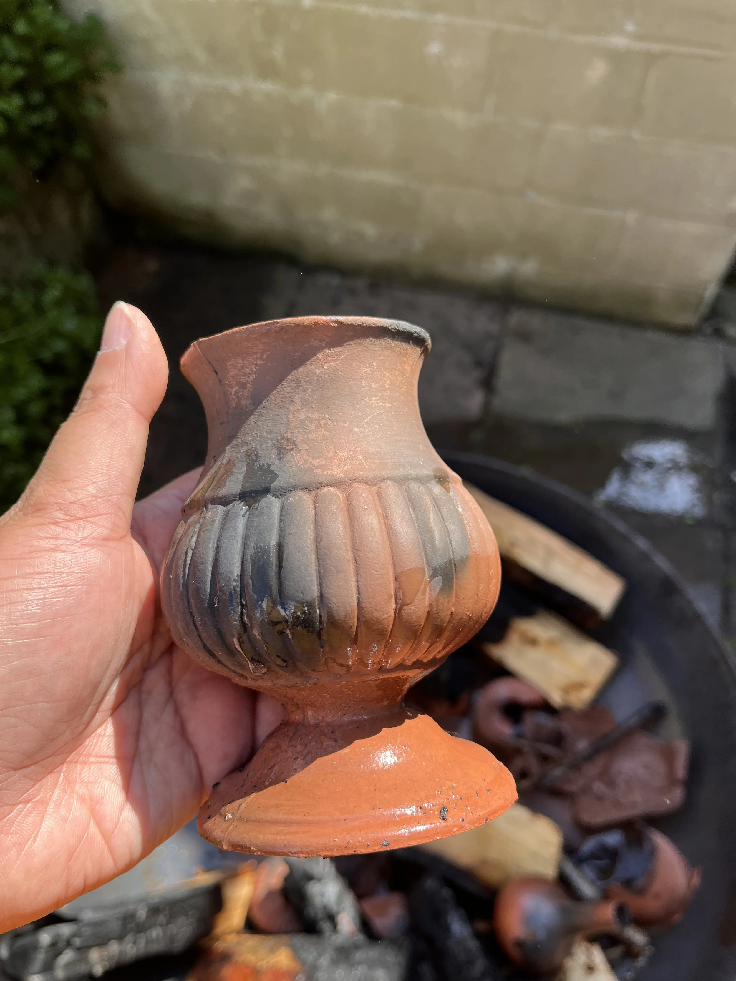 Ani Lacy holding a small, decorative terracotta vase with a ribbed design over a fire pit with logs and other pottery pieces.