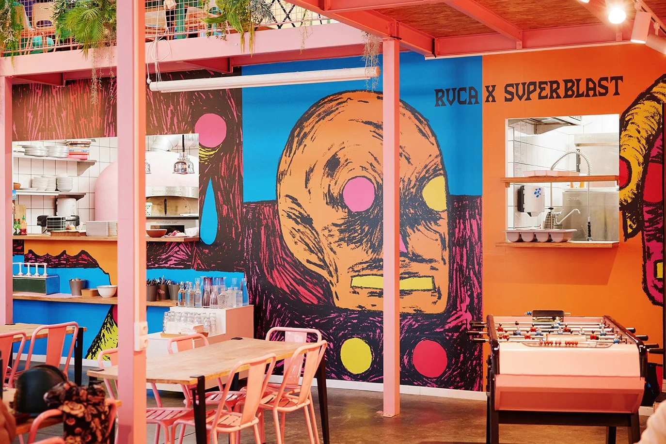 Intérieur coloré d'un café ou d'une salle de jeux, avec un mur peint en style pop art représentant un visage, des tables en bois, des chaises rose, un baby-foot, et une cuisine en arrière-plan avec étagères de vaisselle.