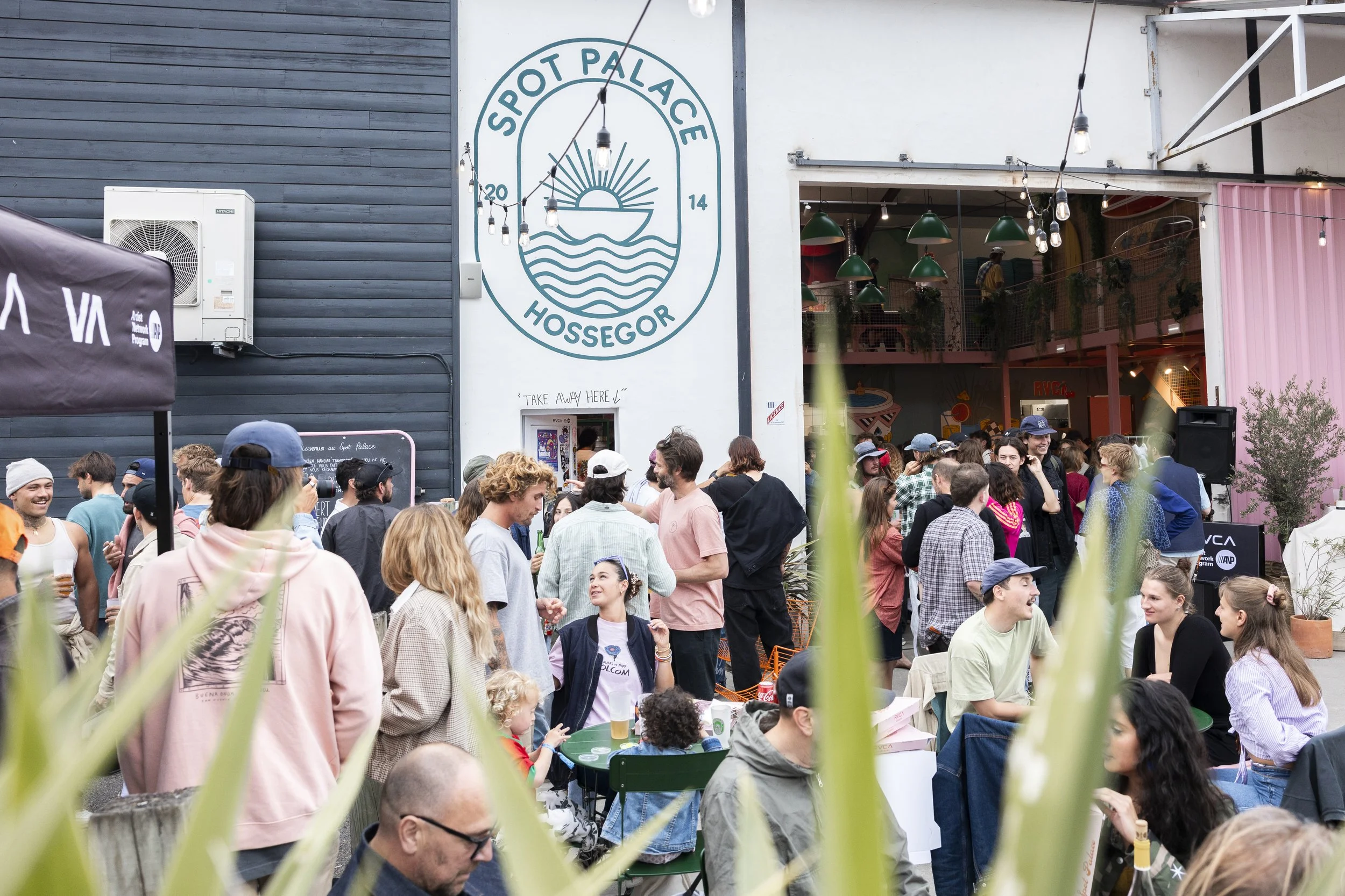 Une grande foule de personnes qui se rassemble à l'extérieur d'un établissement nommé 'Spot Palace Hossegor', avec de la décoration lumineuse suspendue, des tables, des chaises, et des gens qui socialisent, certains avec des boissons à la main.