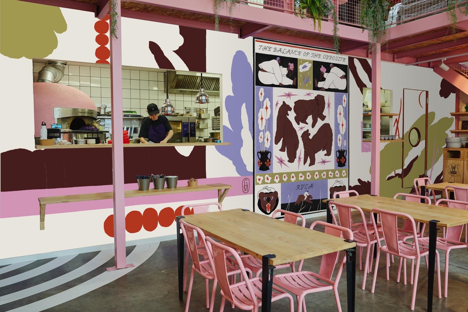 Intérieur d'un café avec murs décorés de peintures colorées, tables en bois et chaises roses, cuisine visible avec chef préparant un plat, et des illustrations de ours et motifs folkloriques.
