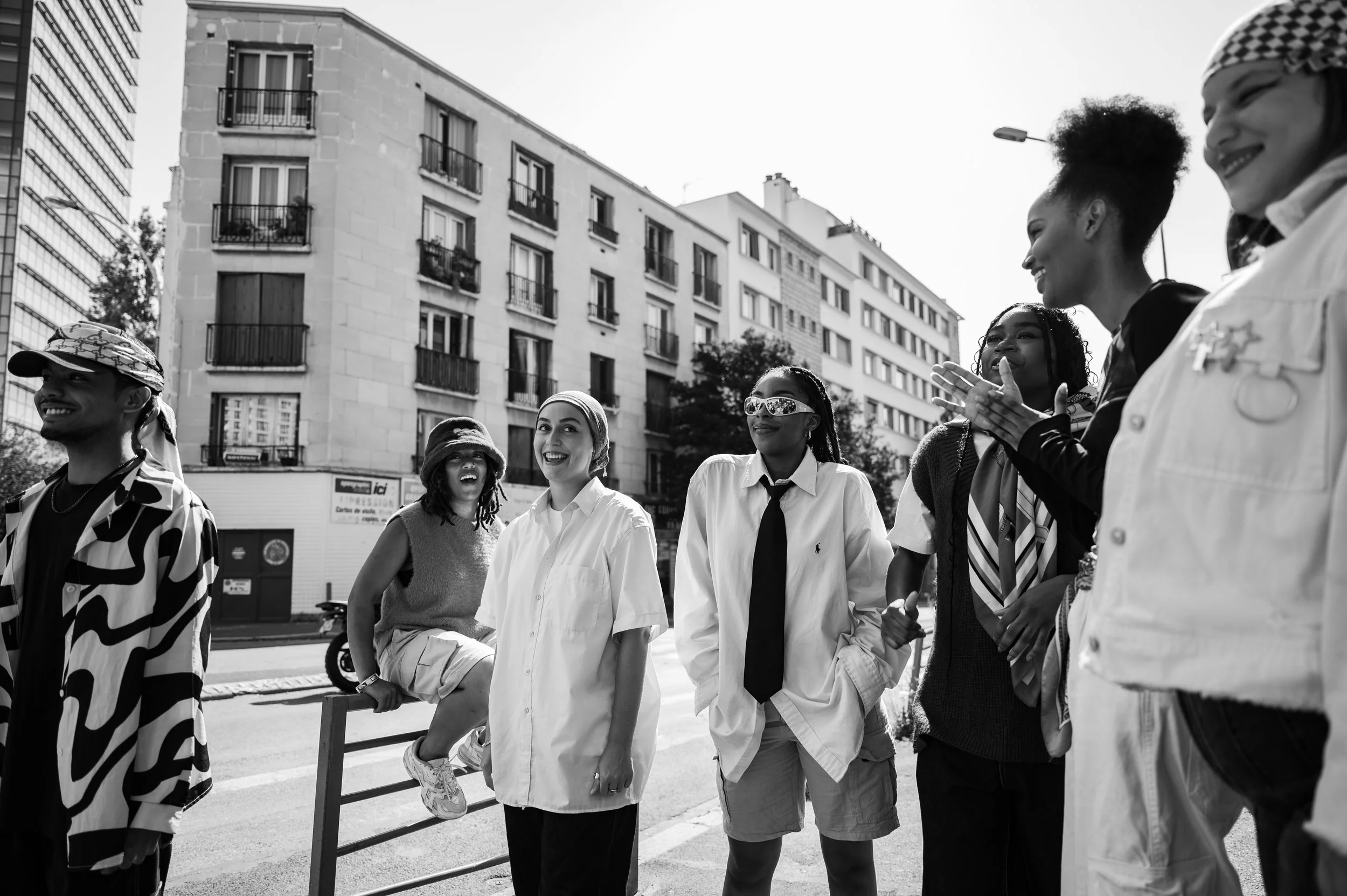 Groupe de personnes diverses souriantes rassemblées au bord de la rue, avec des immeubles résidentiels en arrière-plan, en mode noir et blanc.