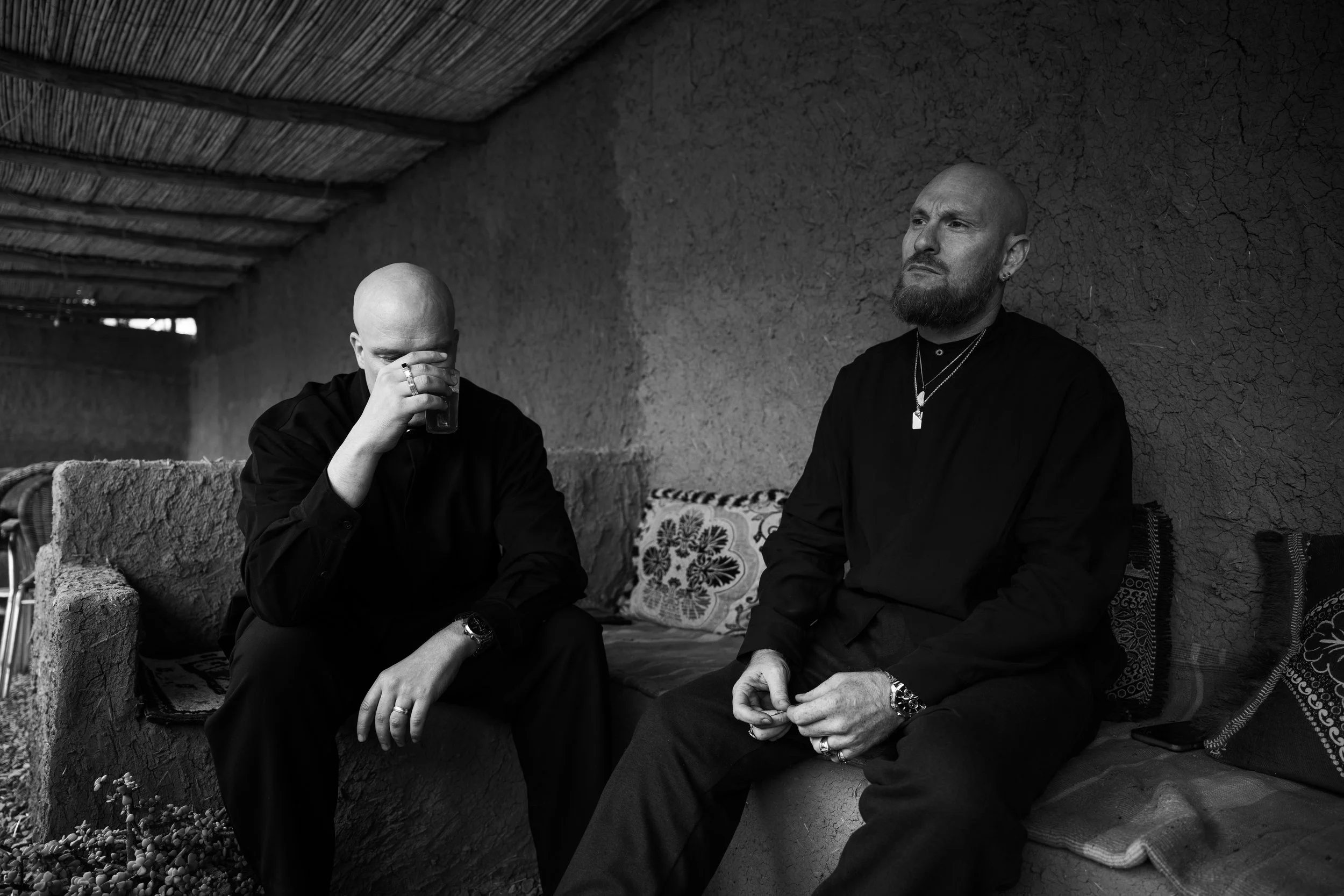 Deux hommes, l'un en train de boire de un verre, assis sur un banc en pierre aucun dans une pièce avec des murs en terre cuite et un plafond en bois, en noir et blanc.