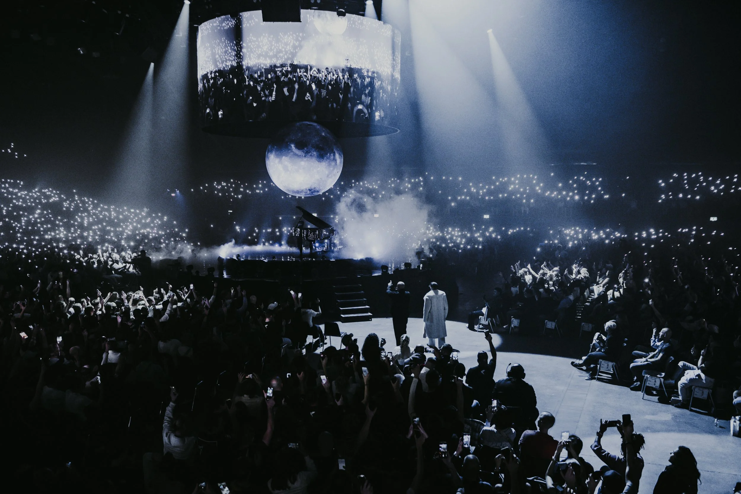 Une scène de concert ou spectacle avec des lumières LED, un écran géant montrant le public, une lune suspendue, et une grande foule assise et debout devant la scène.