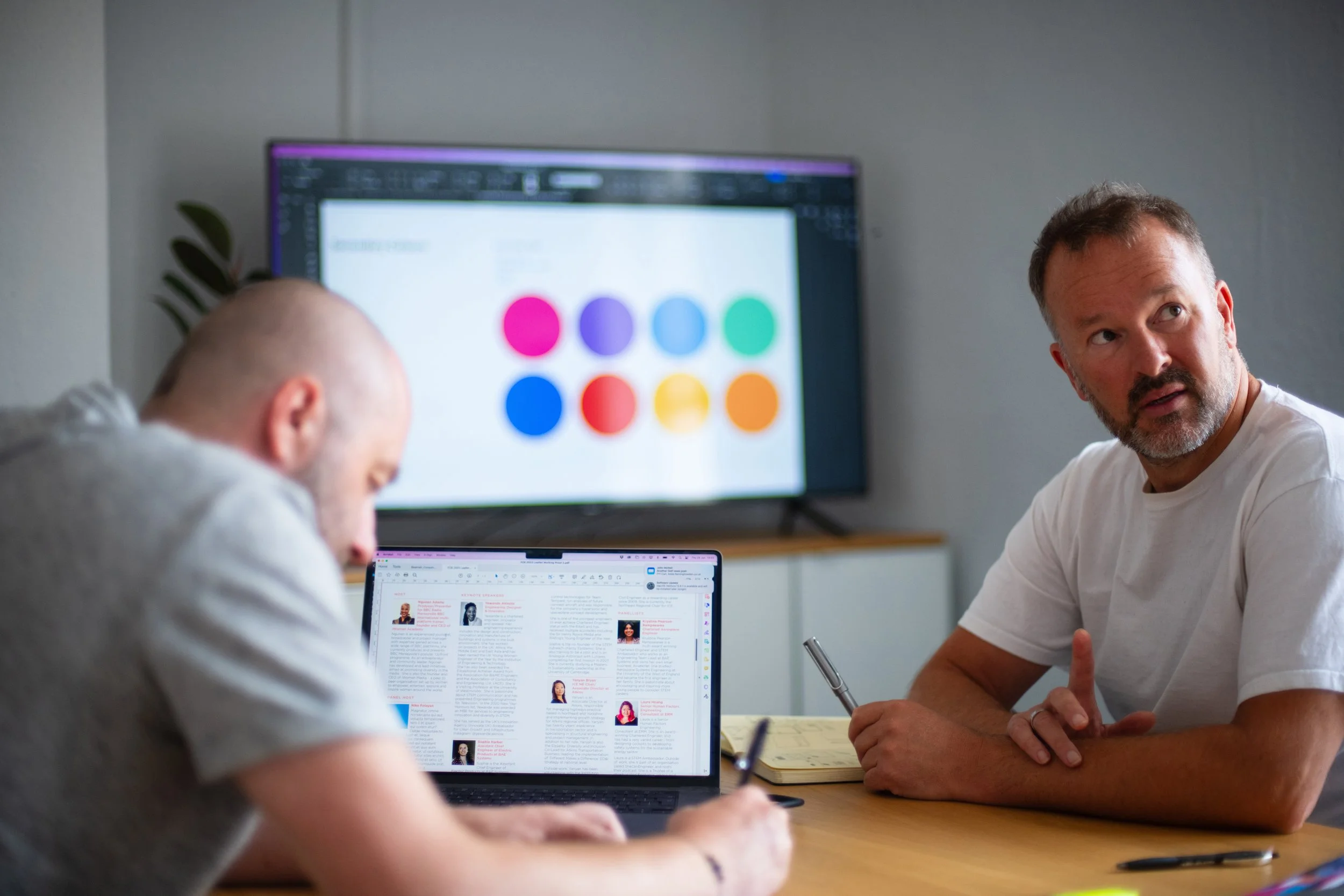 Two men are sitting at a table in a meeting room, one is working on a laptop showing a document with pictures, the other is speaking with a notepad and pen, a large screen behind them displays colorful circles.