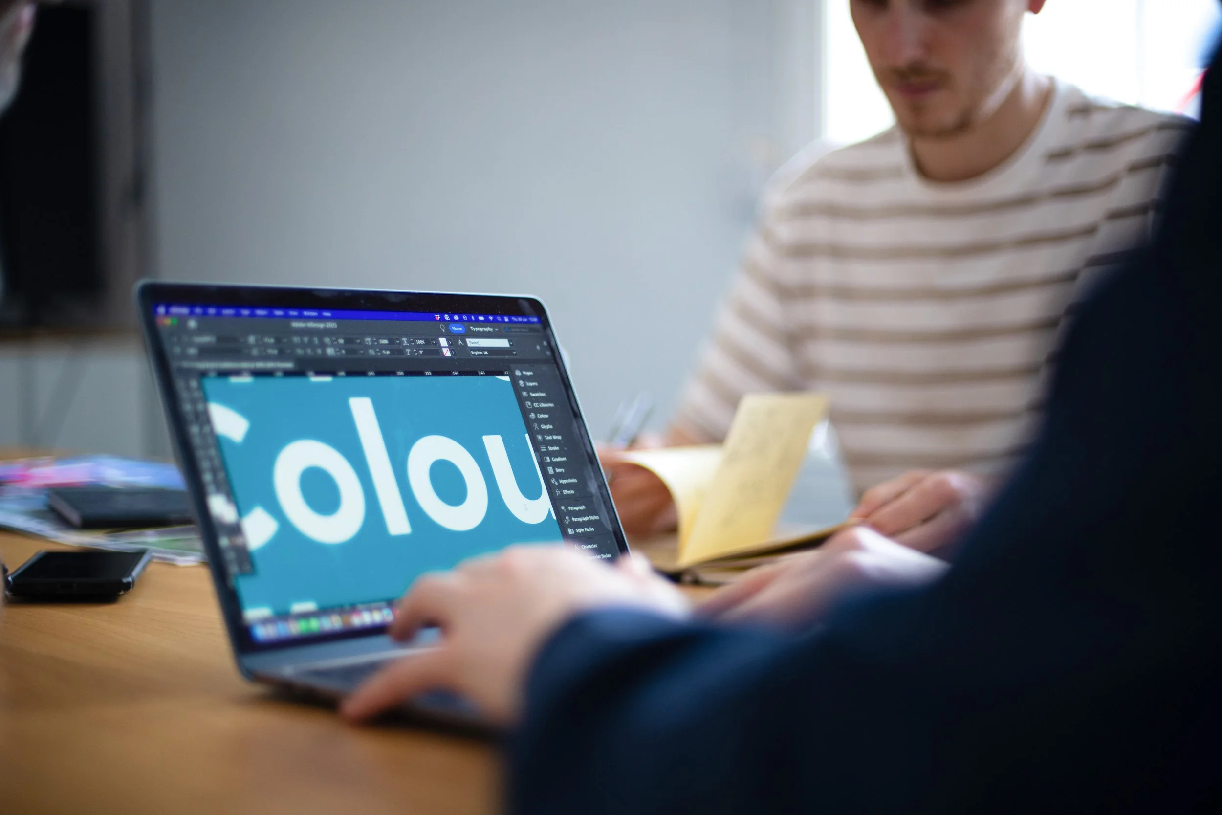 A person using a laptop with a design software open, working on a graphic that features the word 'colour' in large white letters on a blue background.
