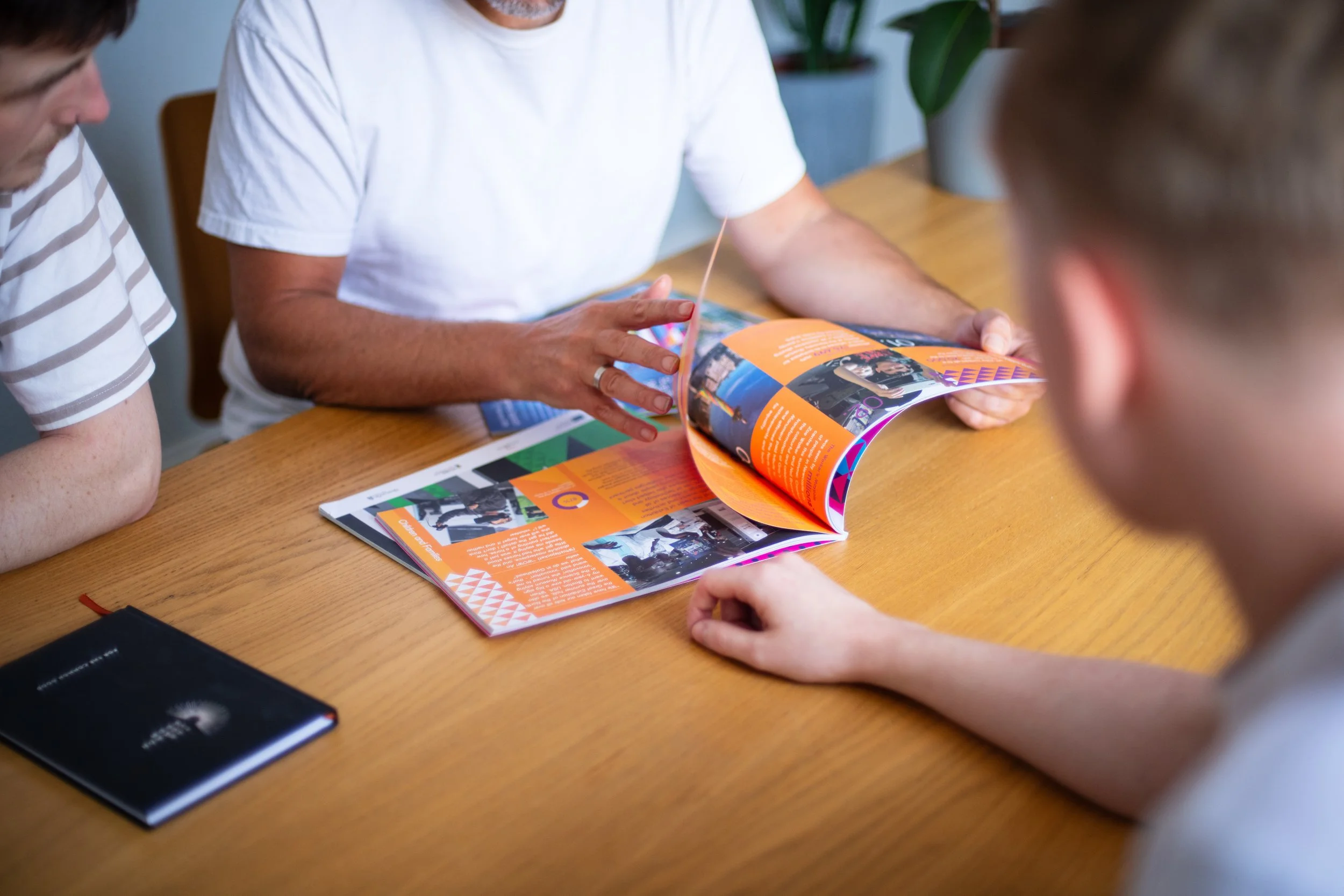 Three men sitting at a wooden table, looking at a colorful magazine. One man in a white t-shirt is turning a page, and the other two are observing.