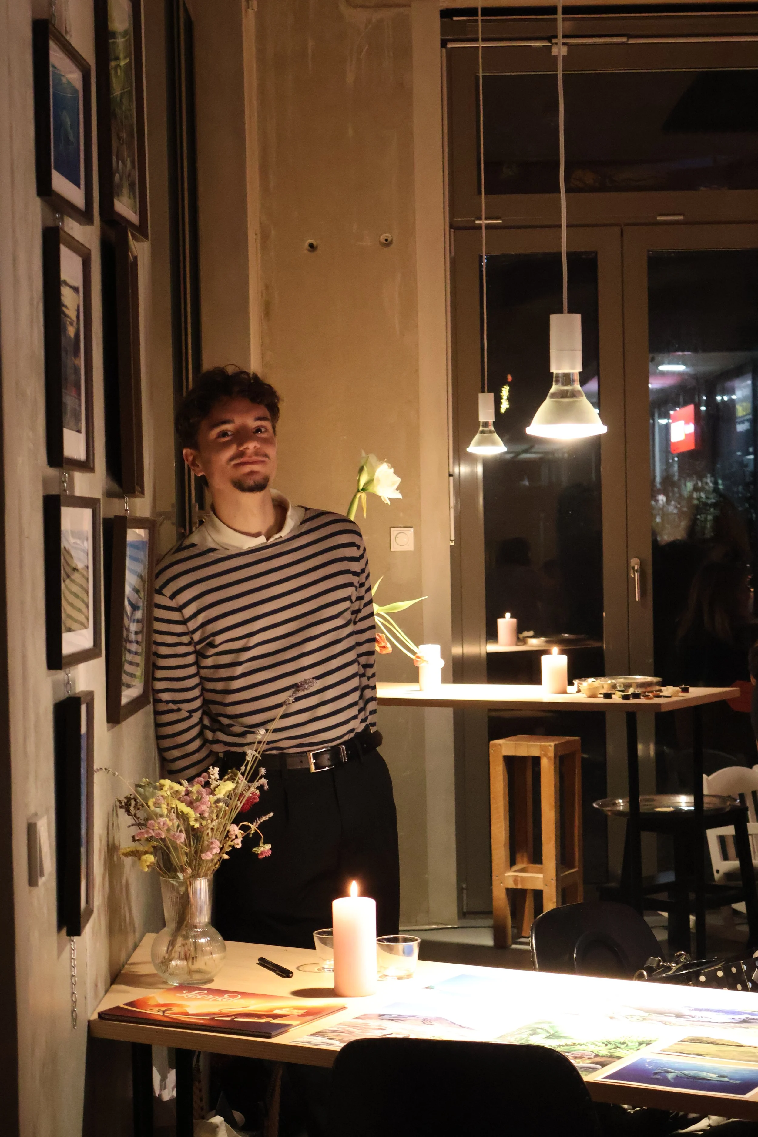 Un homme debout près d'une table avec des bougies allumées et un bouquet de fleurs dans un vase, dans un intérieur chaleureux, décoré avec des cadres sur le mur et des luminaires modernes suspendus au plafond.