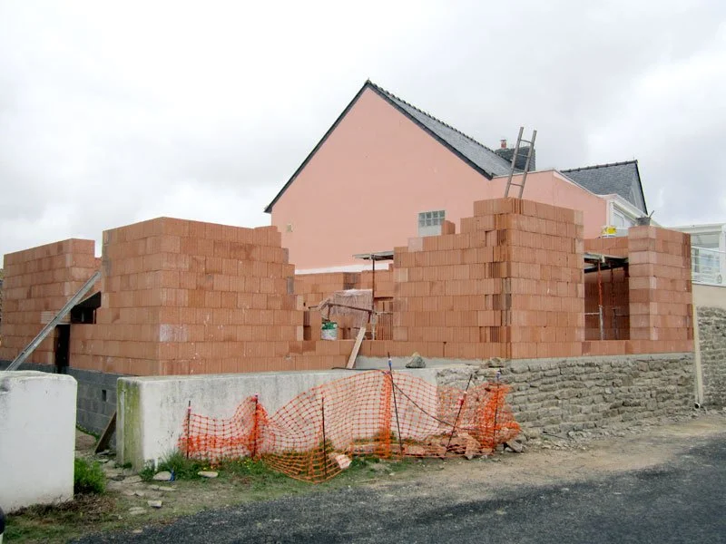 Construction en briques en cours devant une maison rose, avec des protections en filet orange sur le chantier.