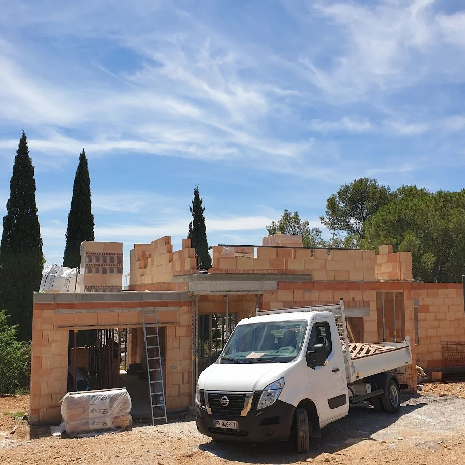 Construction d'une maison avec des briques en terre cuite, un camion blanc à côté, un escalier en aluminium et un ciel bleu avec quelques nuages.