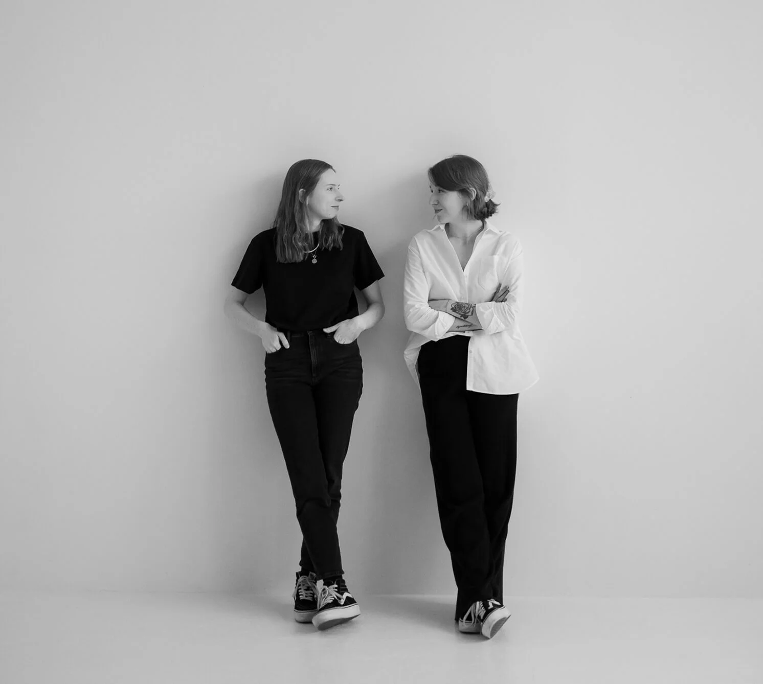 Zwei junge Frauen stehen vor einer weißen Wand, sehen sich an und lächeln. Sie tragen casual Kleidung und Turnschuhe.