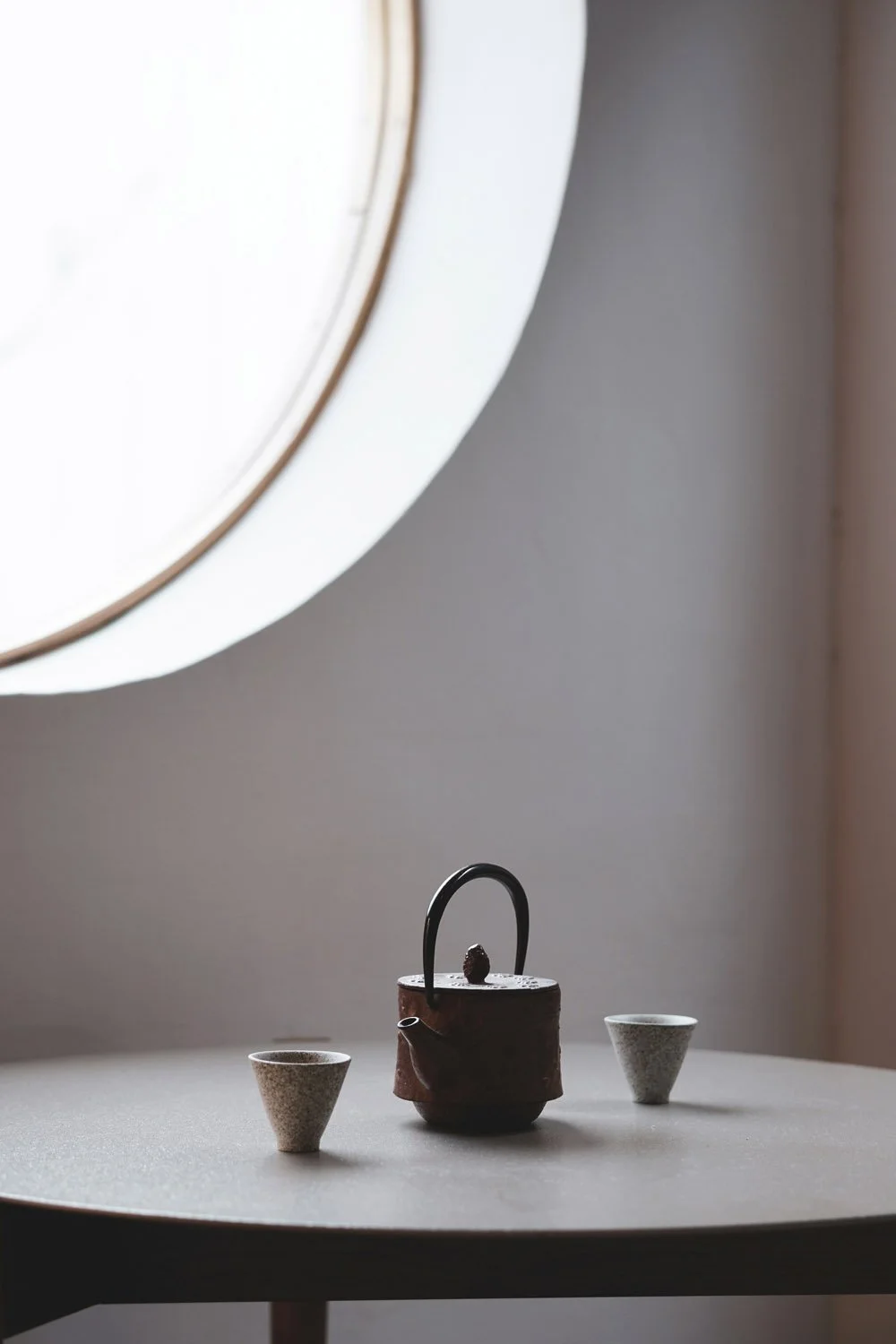 Eine Tasse Tee in einer Teekanne auf einem Tisch mit zwei kleinen Teetassen, im Hintergrund ein rundes Fenster