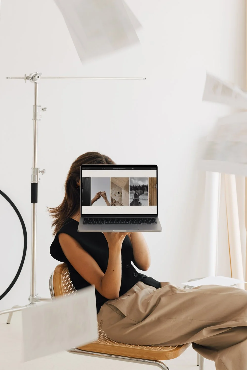 Eine Fotografin sitzt auf einem Stuhl und hält einen Laptop vor das Gesicht, auf dem eine Website mit einer Bilder-Galerie sichtbar ist, in einem hellen Raum mit fotografischer Ausrüstung.