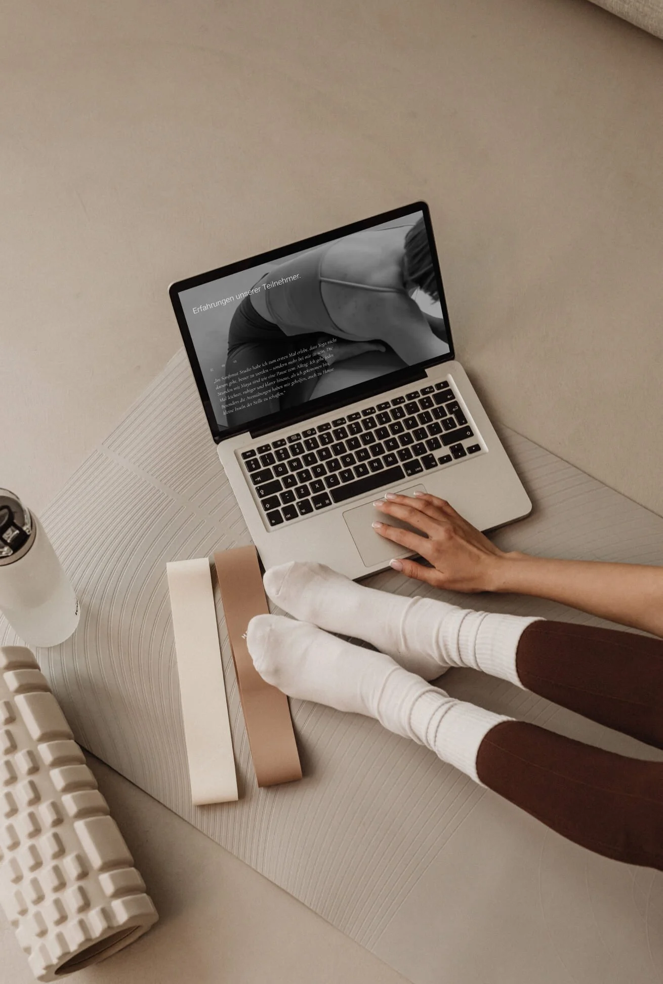Ein Laptop liegt auf einem weißen Tisch, auf dem eine Person mit weißen Socken und braunen Hosen sitzt. Der Laptop zeigt eine Yoga-Übung im Schwarz-Weiß-Bild. Es gibt geometrische Dekorationsstücke und eine wiederrollbare Matte auf dem Tisch.