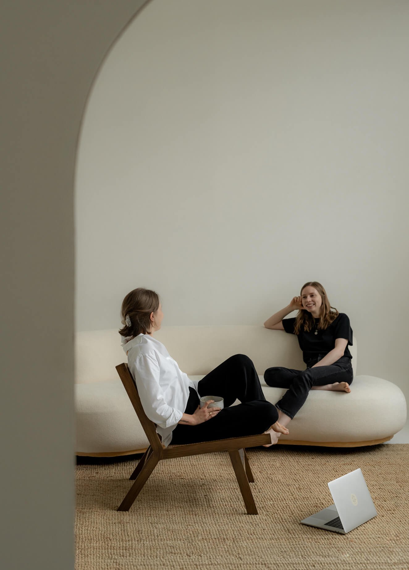 Zwei Frauen sitzen im Gespräch in einem Raum mit minimalistischem Design, einer auf einem Holzstuhl und die andere auf einer großen weißen Couch, mit einem Laptop auf dem Boden.