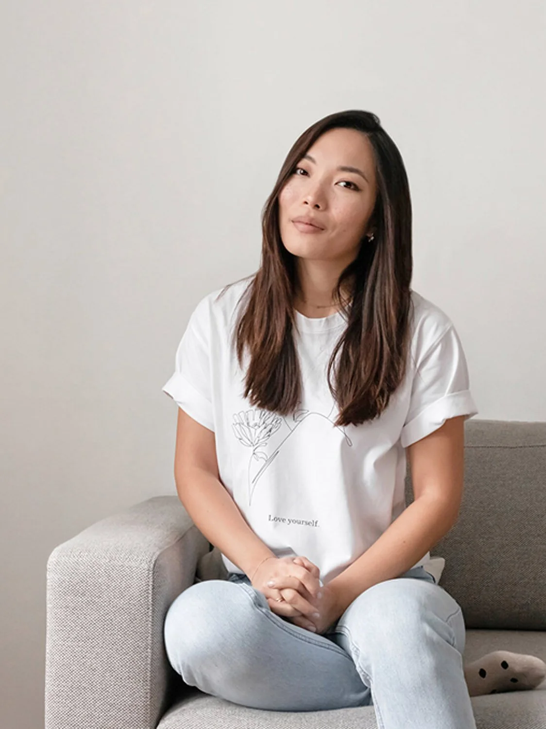 Eine Frau sitzt auf einem grauen Sofa vor einer weißen Wand, trägt ein weißes T-Shirt mit einem Blumen-Design undText, und blickt freundlich in die Kamera.