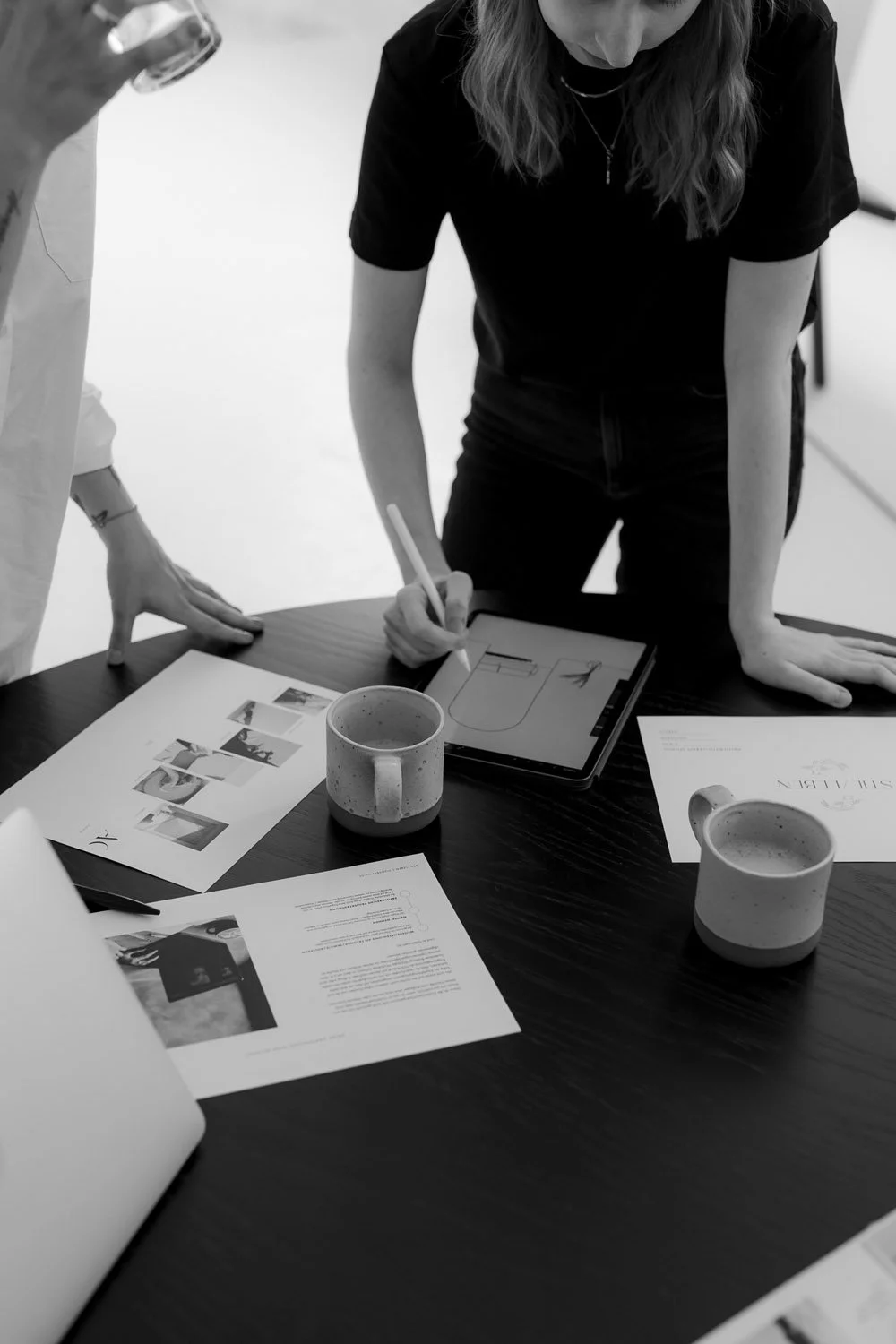 Zwei Frauen arbeiten an einem Tisch mit Dokumenten, einem Tablet, einem Stift und zwei Tassen Kaffee in einem Büro oder Besprechungsraum.