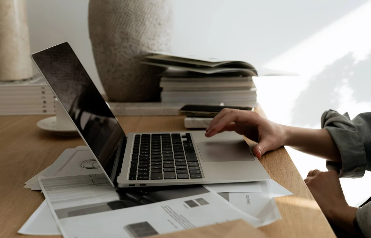Person arbeitet an einem Laptop auf einem Holztisch mit Papieren, Büchern und einem großen Keramikvase im Hintergrund.