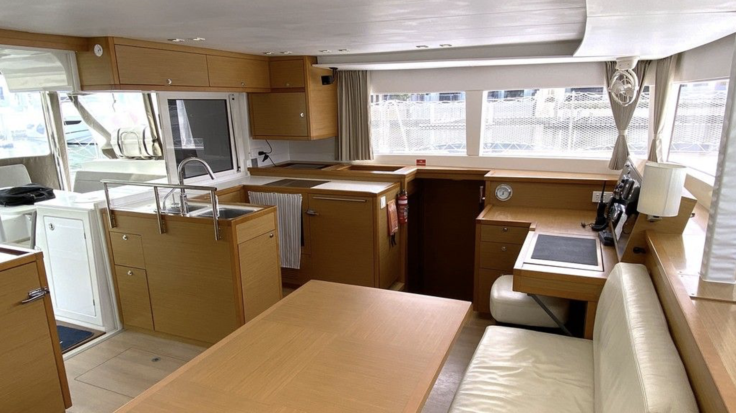 Innenaufnahme einer Yacht-Küche mit Holzschränken, einer Spüle, einem kleinen Esstisch, einem Fenster mit Vorhängen und verschiedenen Geräten.
