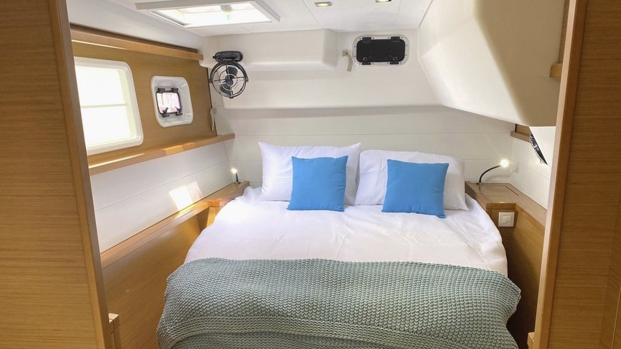 Ein Schlafraum mit einem Doppelbett, vier Kissen (zwei weiße, zwei blaue), kleinen Wandlampen, zwei Fenster, einem Ventilator und einer kleinen Luke in der Decke, alles in Holz- und Weißtönen gehalten.