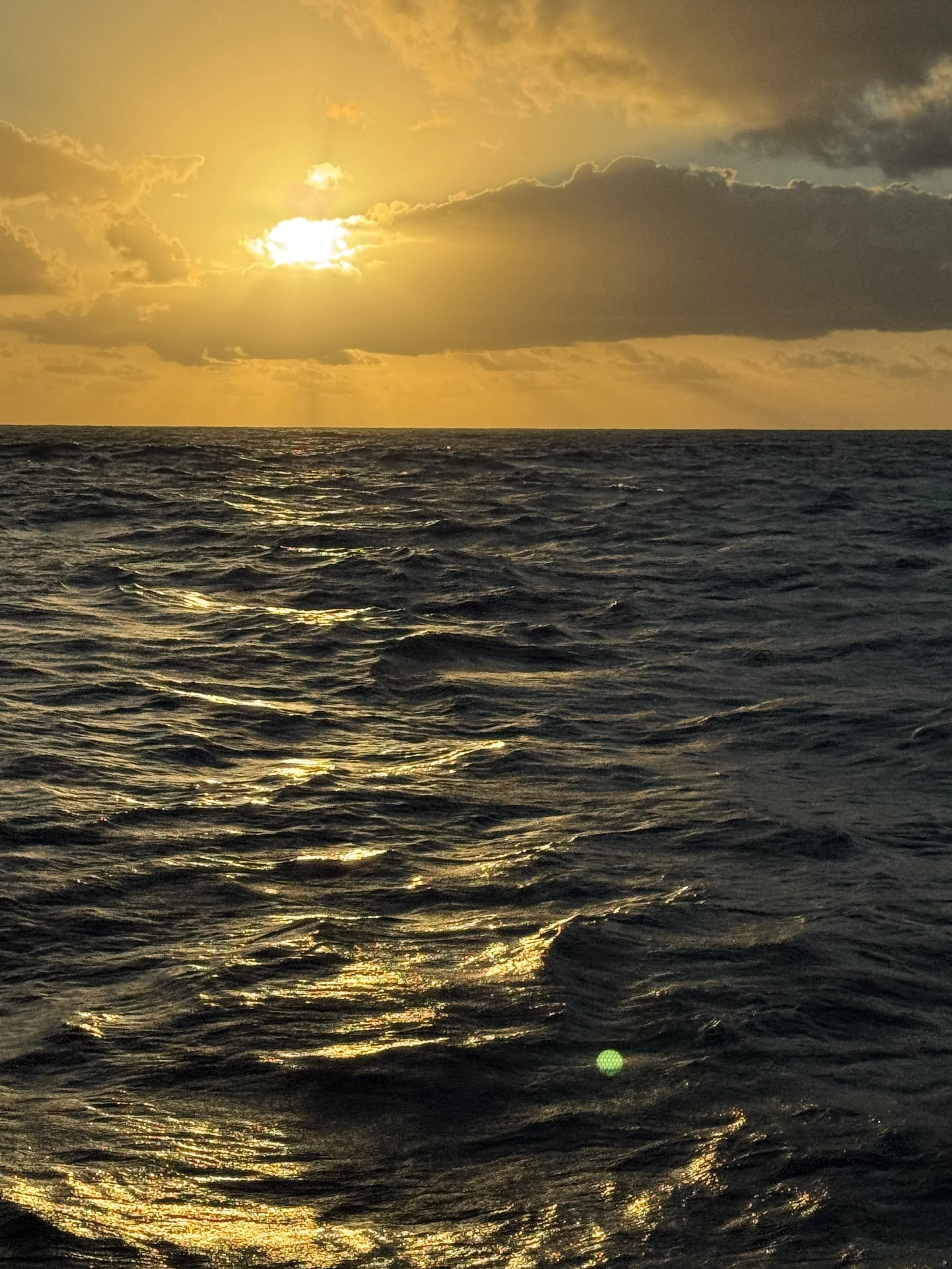 Ein Meer bei Sonnenuntergang mit dunklen Wolken im Himmel, das Sonnenlicht spiegelt sich auf den Wellen.