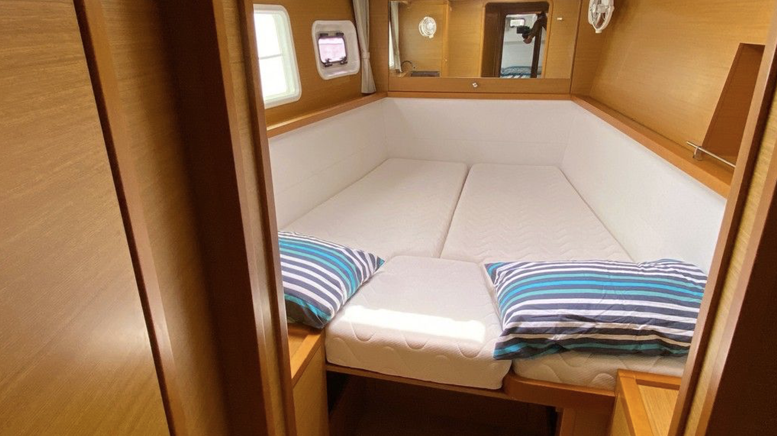 Innenraum eines Bootes mit einem L-förmigen Bett, weißen Bettdecken, zwei Kissen mit blauen und schwarzen Streifen, Holzpaneelen und kleinen Fenstern.