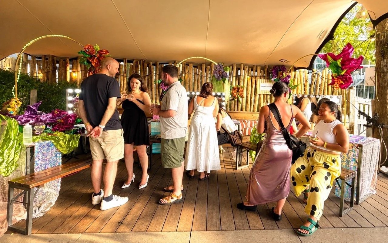 People socializing at a decorated outdoor party with colorful flowers, string lights, and a wooden fence background.