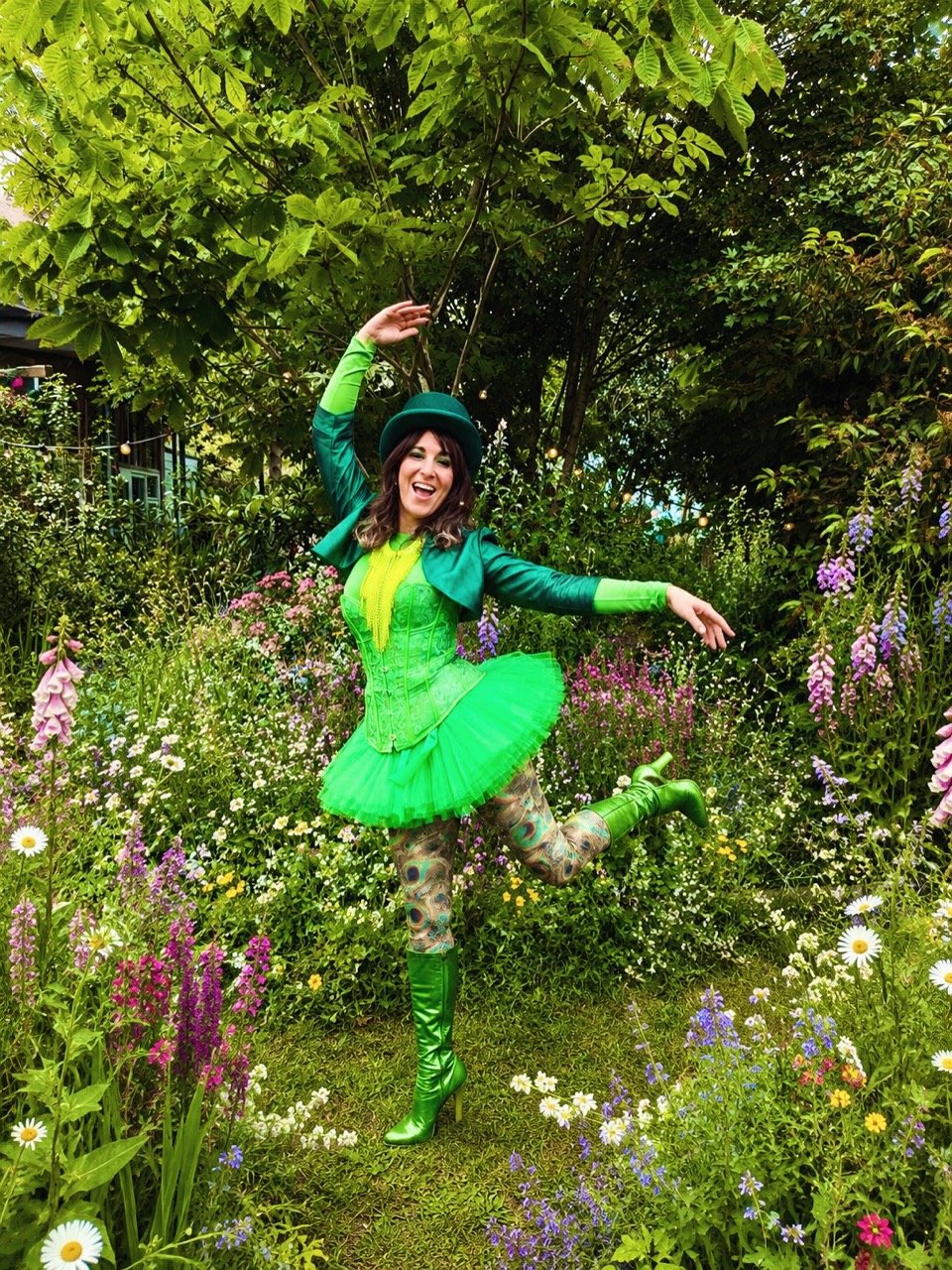 A woman in a vibrant green costume dancing in a garden filled with colorful flowers and lush green trees.