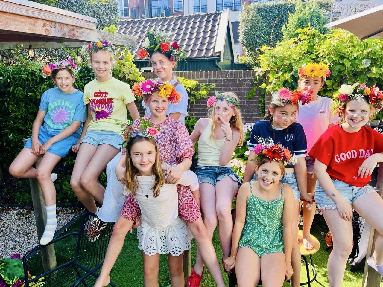 A group of ten young girls wearing colorful shirts and shorts with flower crowns on their heads, gathered in a garden with trees and buildings in the background, smiling and posing for a photo.