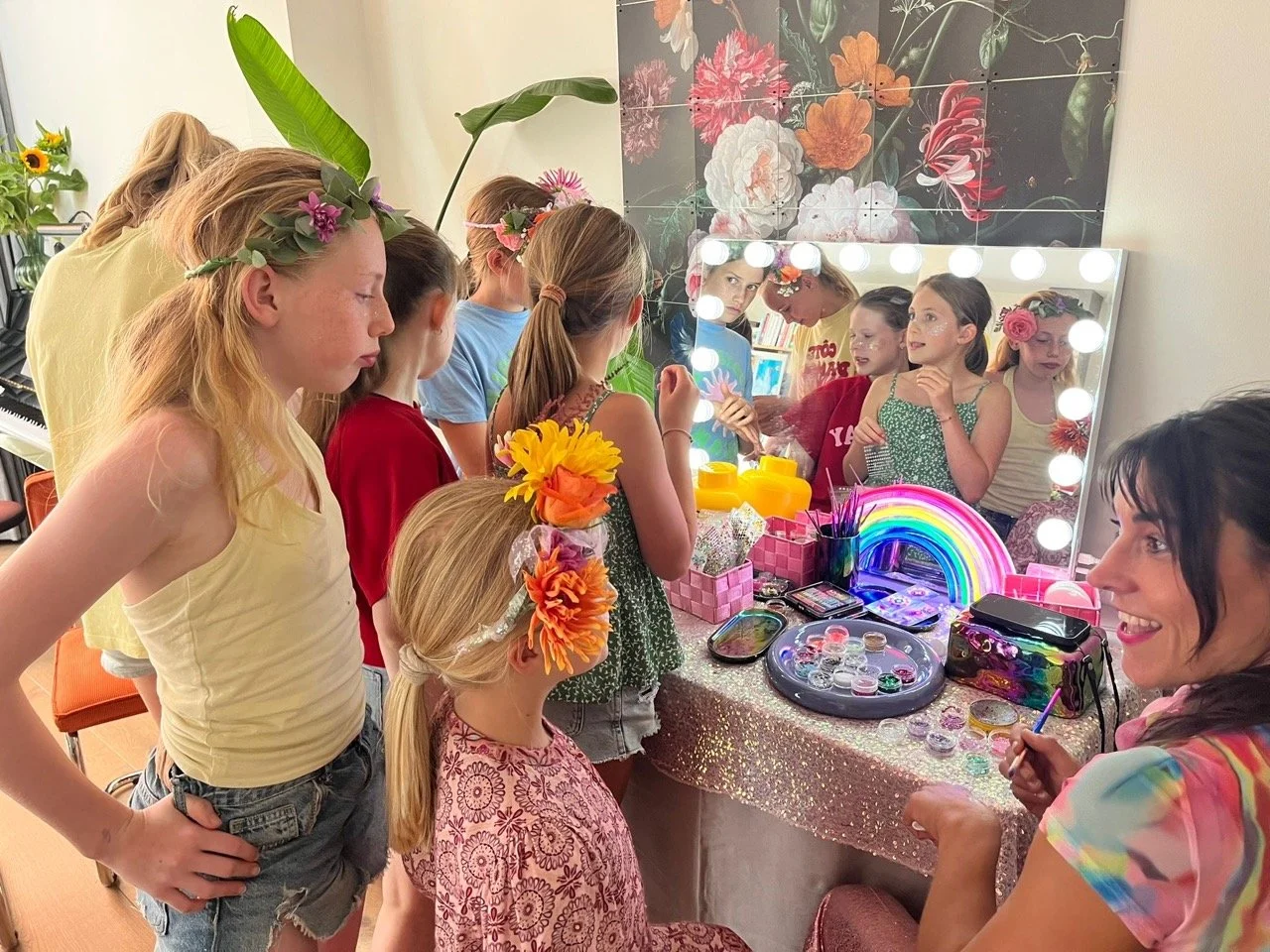 Group of girls with floral headbands gathered around a vanity mirror decorated with lights and rainbow decor, in a room with floral wall art and plants, preparing for a celebration.