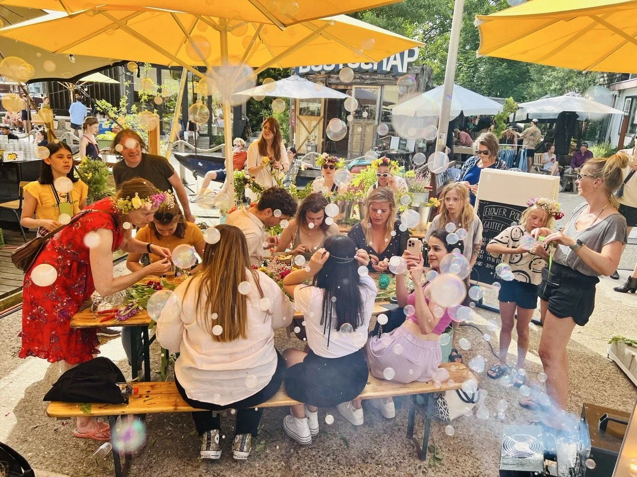 People gathered at an outdoor flower-making event under yellow umbrellas, crafting floral arrangements with various tools and materials.