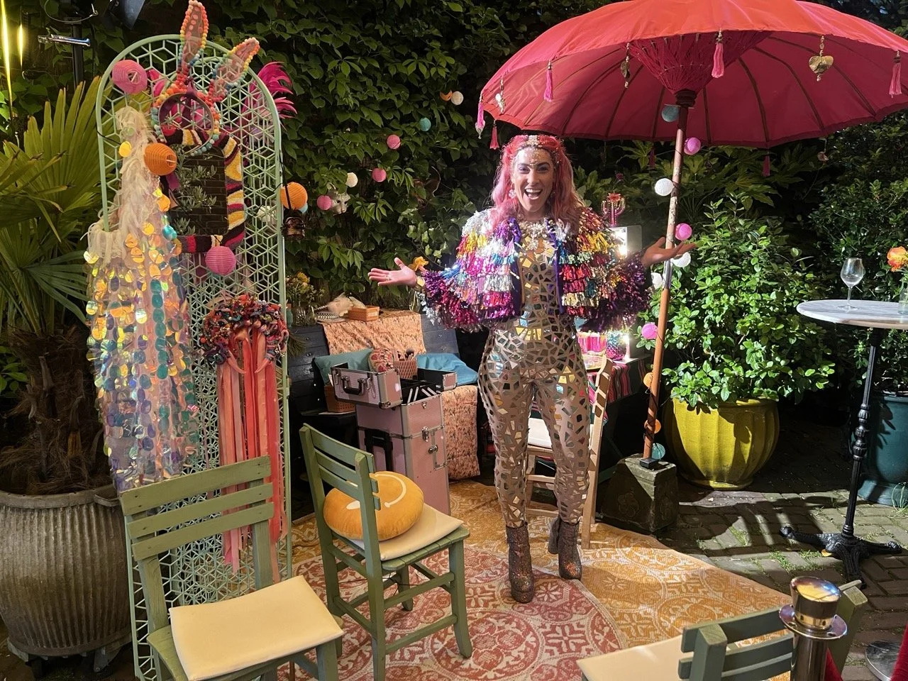Woman smiling at a vibrant outdoor party with colorful decorations, lampions, and a pink umbrella, surrounded by plants and eclectic furniture.