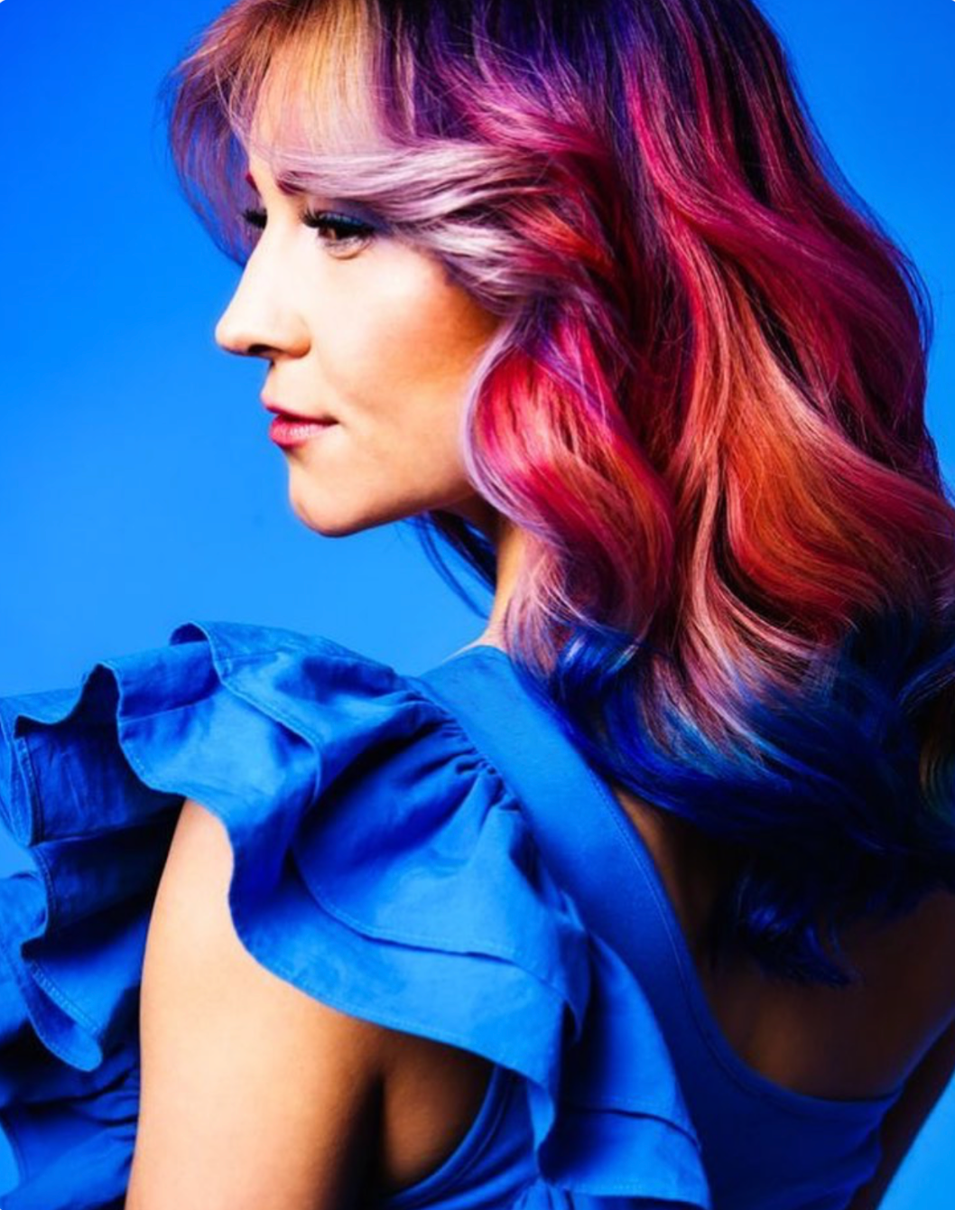 A woman with colorful, wavy hair in shades of pink, purple, and blue, wearing a blue ruffled top, against a blue background.