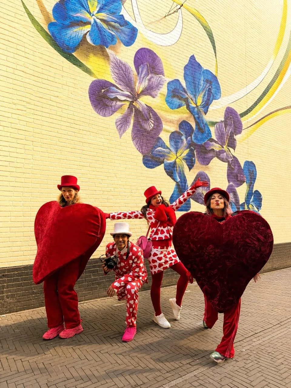 Four women dressed in red and white heart-themed costumes and red hats, holding oversized heart-shaped props, standing in front of a yellow brick wall with a colorful floral mural.