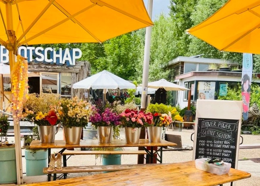 Outdoor flower booth with colorful flowers in metal buckets, yellow umbrellas, and a chalkboard sign, surrounded by green trees and other market stalls.